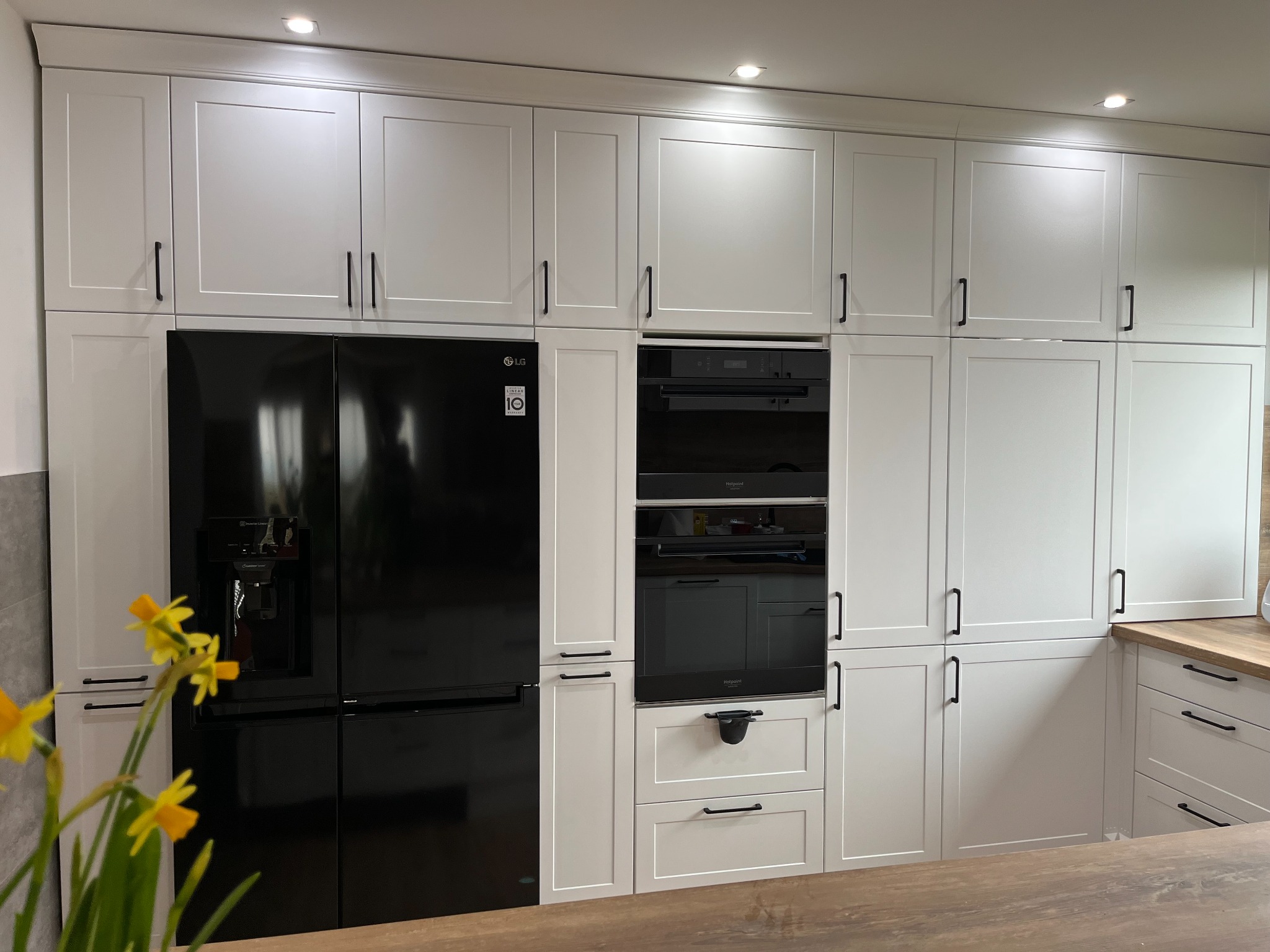 Biała zabudowa kuchenna z czarnym sprzętem AGD: lodówka side-by-side i piekarnik w zabudowie, uzupełniona drewnianym blatem i bukietem żółtych narcyzów w pierwszym planie.