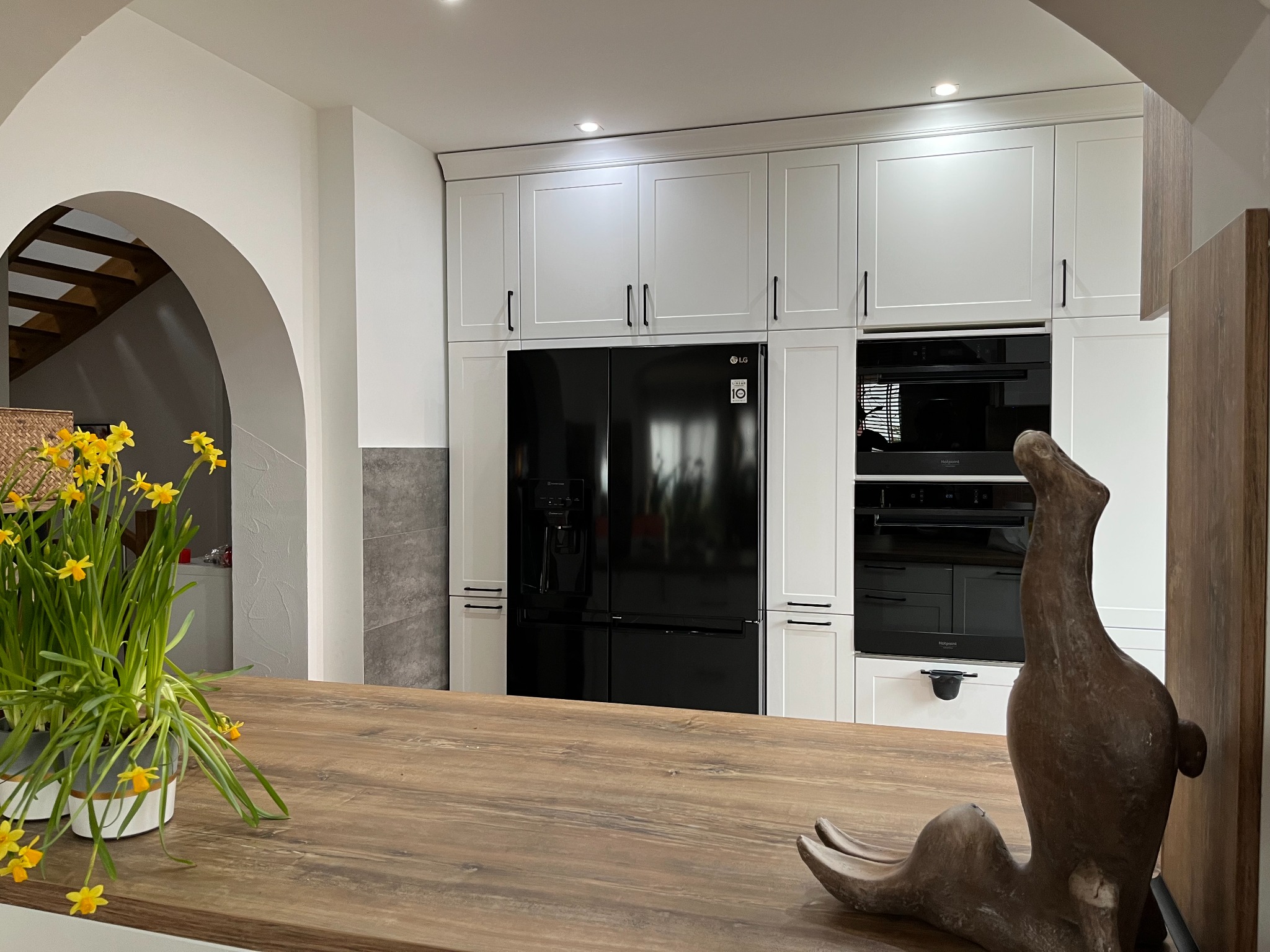 Nowoczesna kuchnia z białymi szafkami, czarną lodówką LG i drewnianym blatem, na którym stoi doniczka z żółtymi narcyzami i drewniana rzeźba.