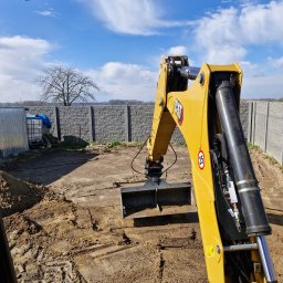 Marcin Wojciech - Żółta koparka CAT na placu budowy, widok z kabiny operatora, w tle betonowy mur i błękitne niebo z chmurami. Ślady opon na ziemi.