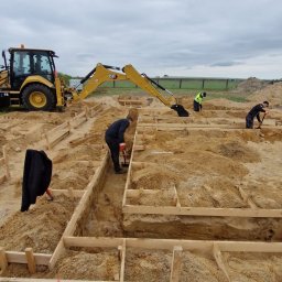 Marcin Wojciech - Żółta koparko-ładowarka CAT i pracownicy przygotowujący drewniany szalunek pod fundamenty na piaszczystym terenie. Jeden z pracowników używa ubijaka.