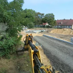 Widok z kabiny żółtej koparki na plac budowy z przygotowanym podłożem z kruszywa, widoczne fundamenty w oddali i budynki mieszkalne w tle.
