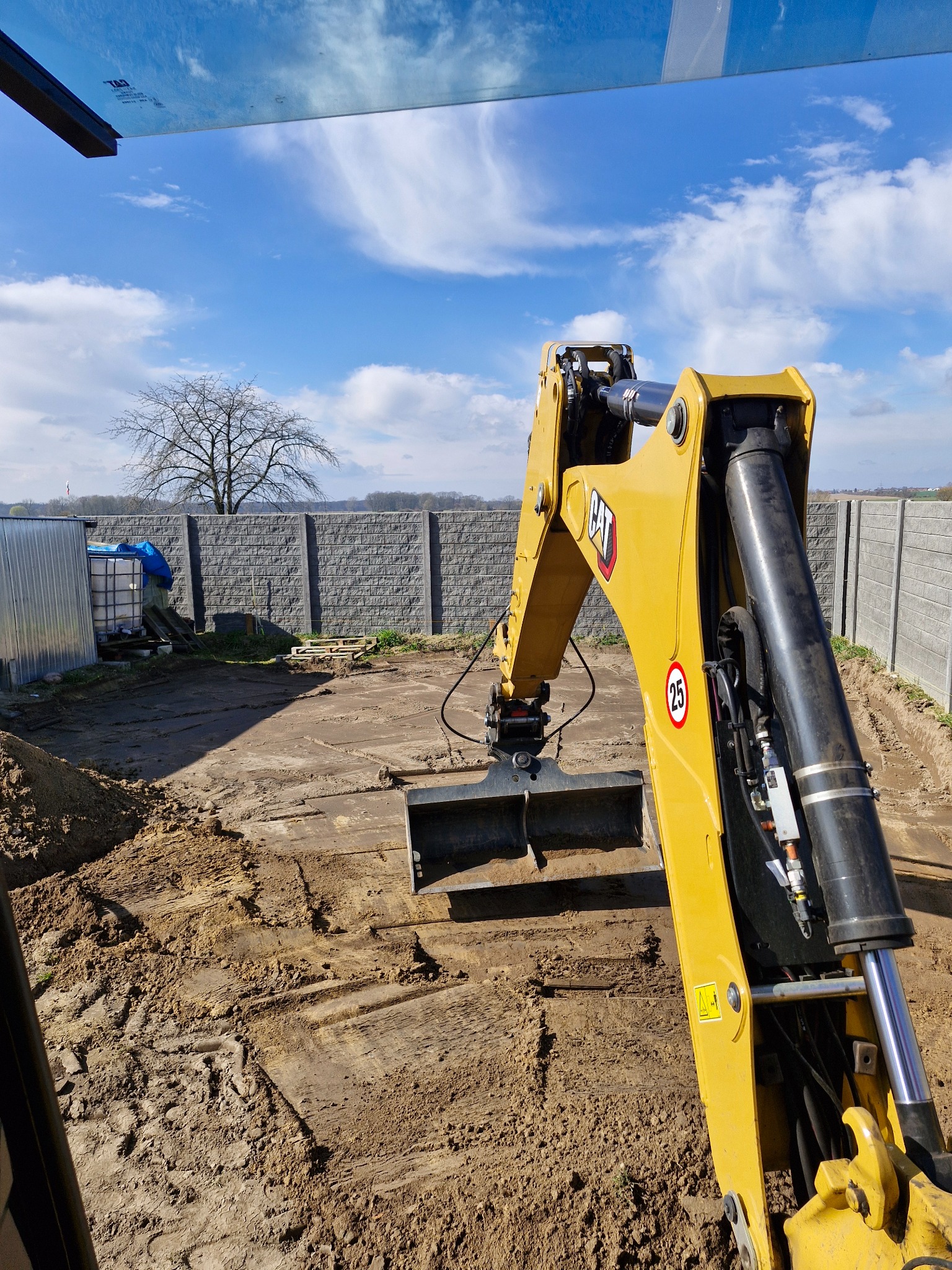 Żółta koparka CAT na placu budowy, widok z kabiny operatora, w tle betonowy mur i błękitne niebo z chmurami. Ślady opon na ziemi.