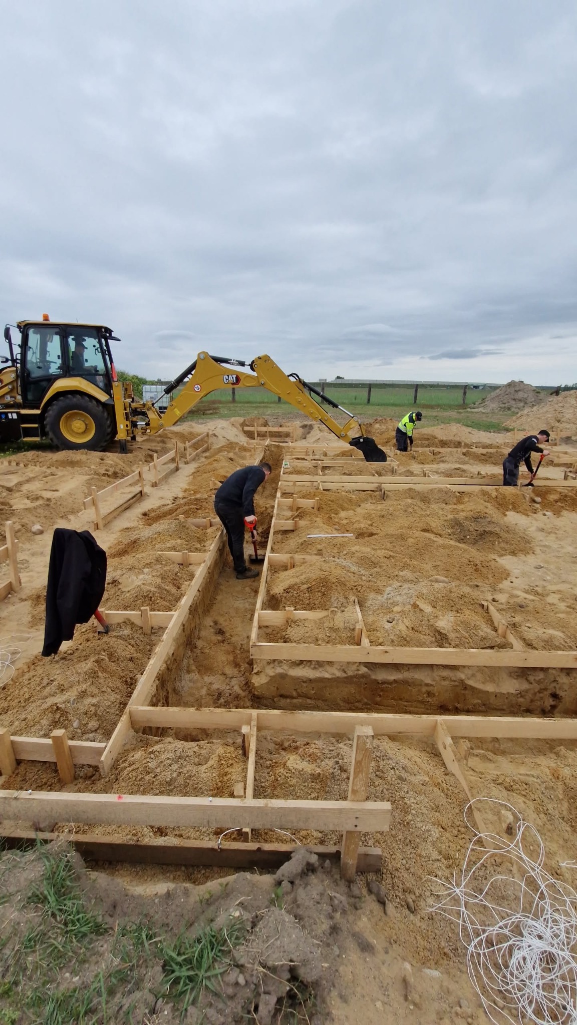 Żółta koparko-ładowarka CAT i pracownicy przygotowujący drewniany szalunek pod fundamenty na piaszczystym terenie. Jeden z pracowników używa ubijaka.