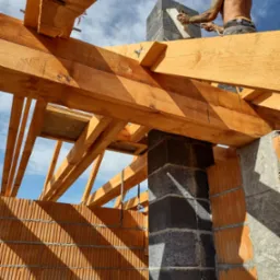 Murarz z tatuażami wyrównuje zaprawę na kominie z szarych bloczków, widoczny z perspektywy wnętrza budynku w trakcie budowy dachu z drewnianych belek.