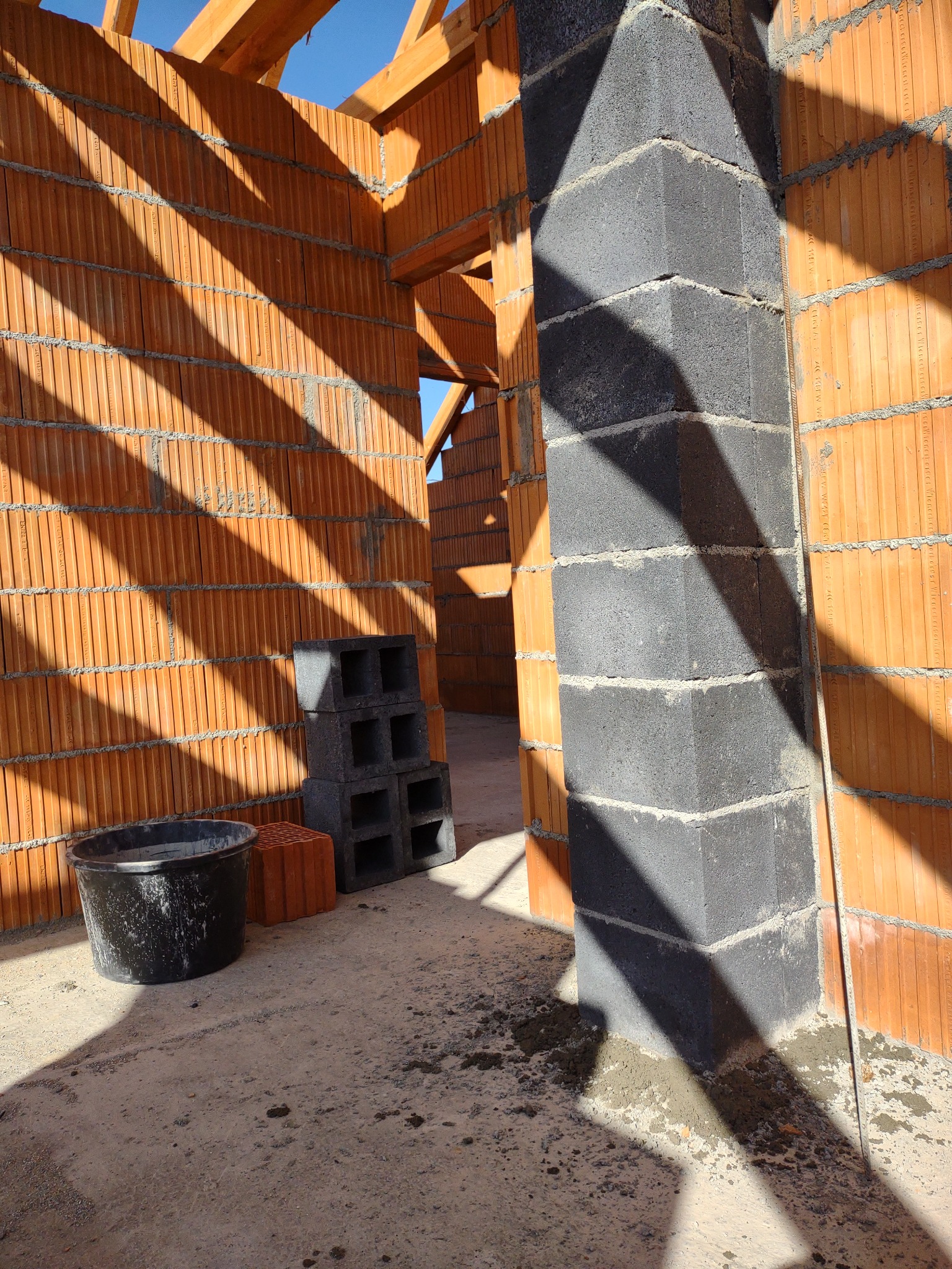 Wnętrze w trakcie budowy: ściany z pomarańczowych pustaków ceramicznych, szare bloczki betonowe ułożone w kolumnę, konstrukcja drewniana dachu, wiadro i cień.