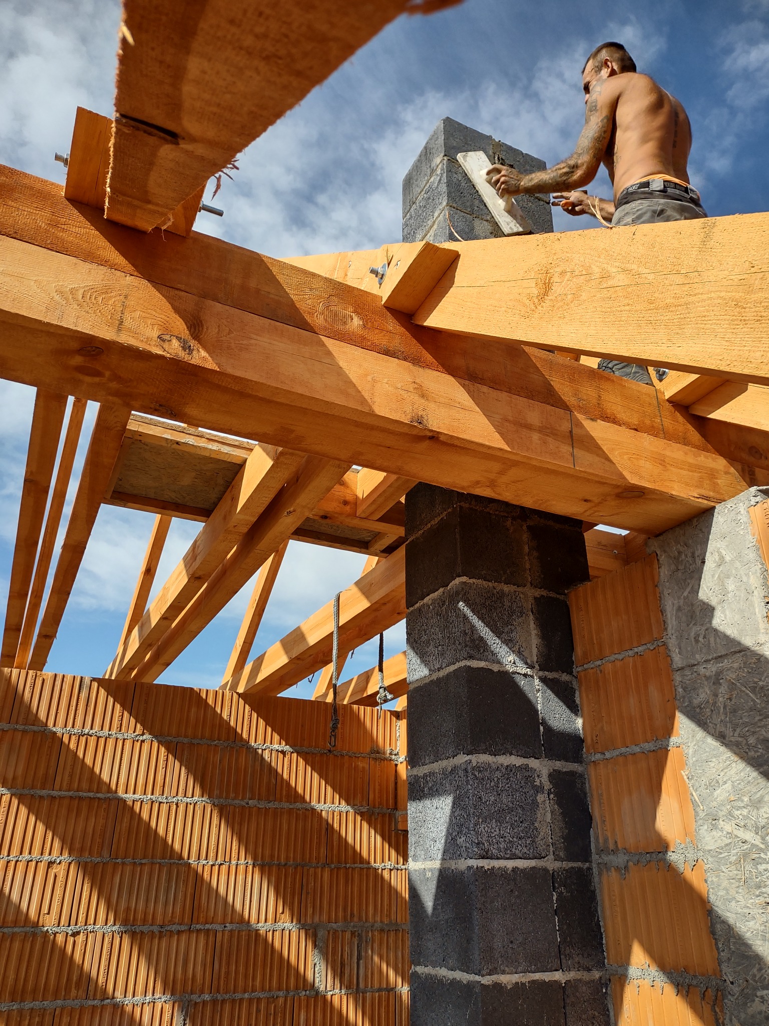 Murarz z tatuażami wyrównuje zaprawę na kominie z szarych bloczków, widoczny z perspektywy wnętrza budynku w trakcie budowy dachu z drewnianych belek.