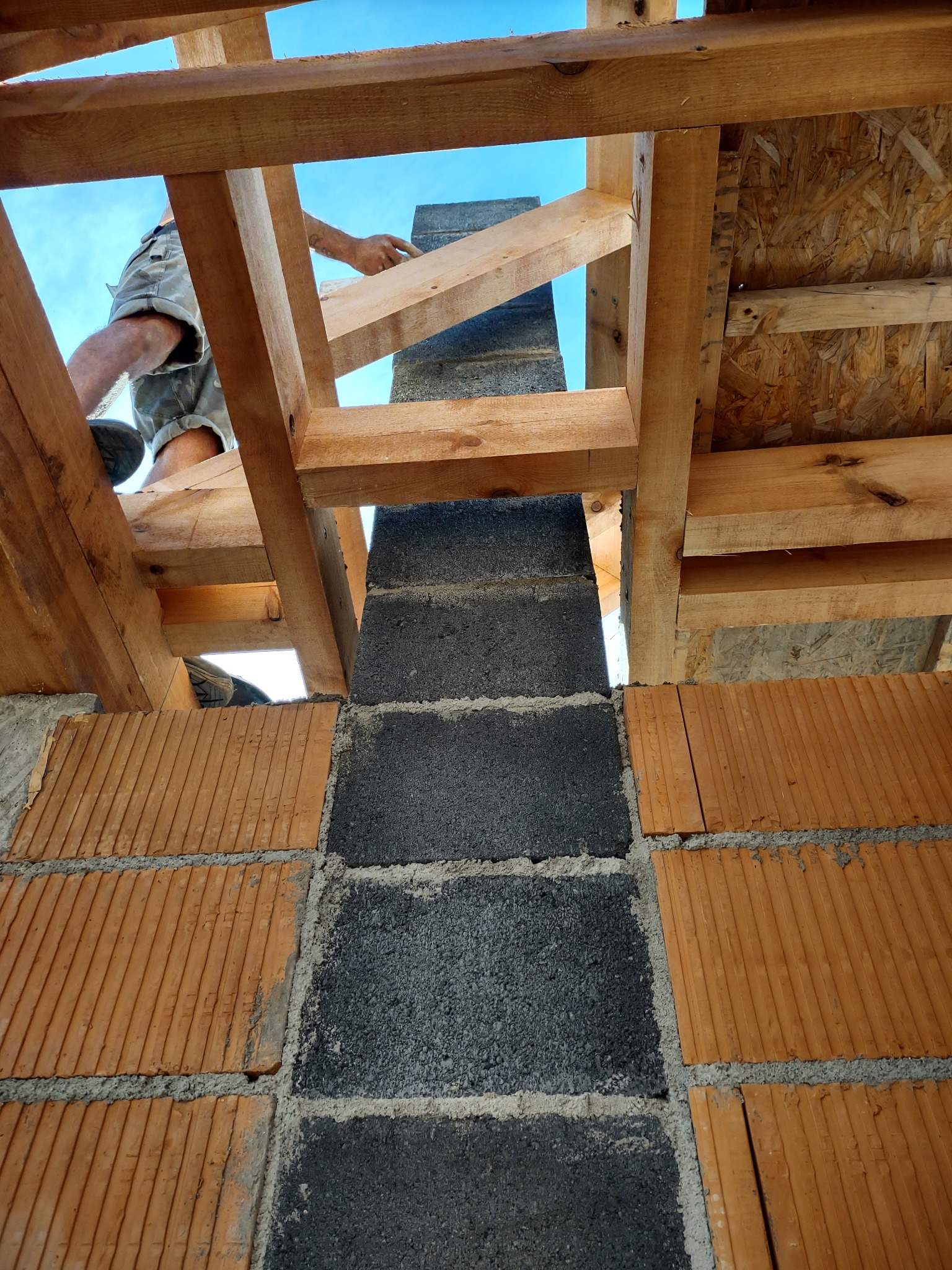 Wznoszenie komina z szarych bloczków betonowych, widok od dołu na tle drewnianej konstrukcji dachu i fragmentu niebieskiego nieba.