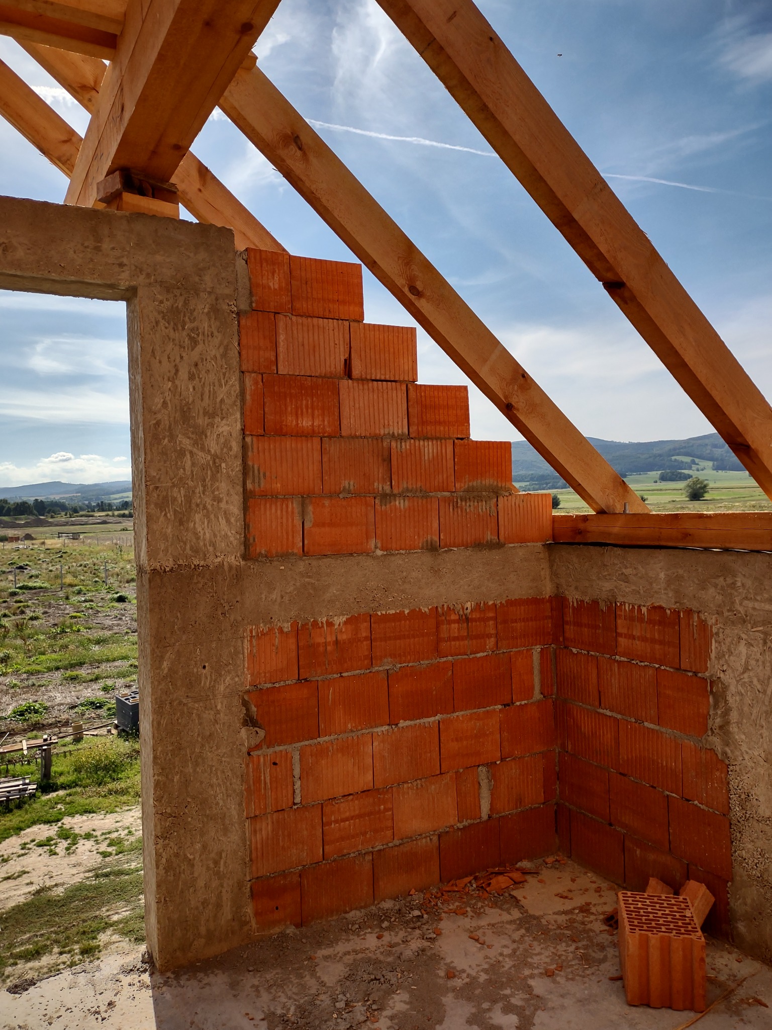 Wznoszenie muru z czerwonej cegły ceramicznej na zaprawie, widoczne drewniane elementy konstrukcji dachu i fragment krajobrazu w tle.