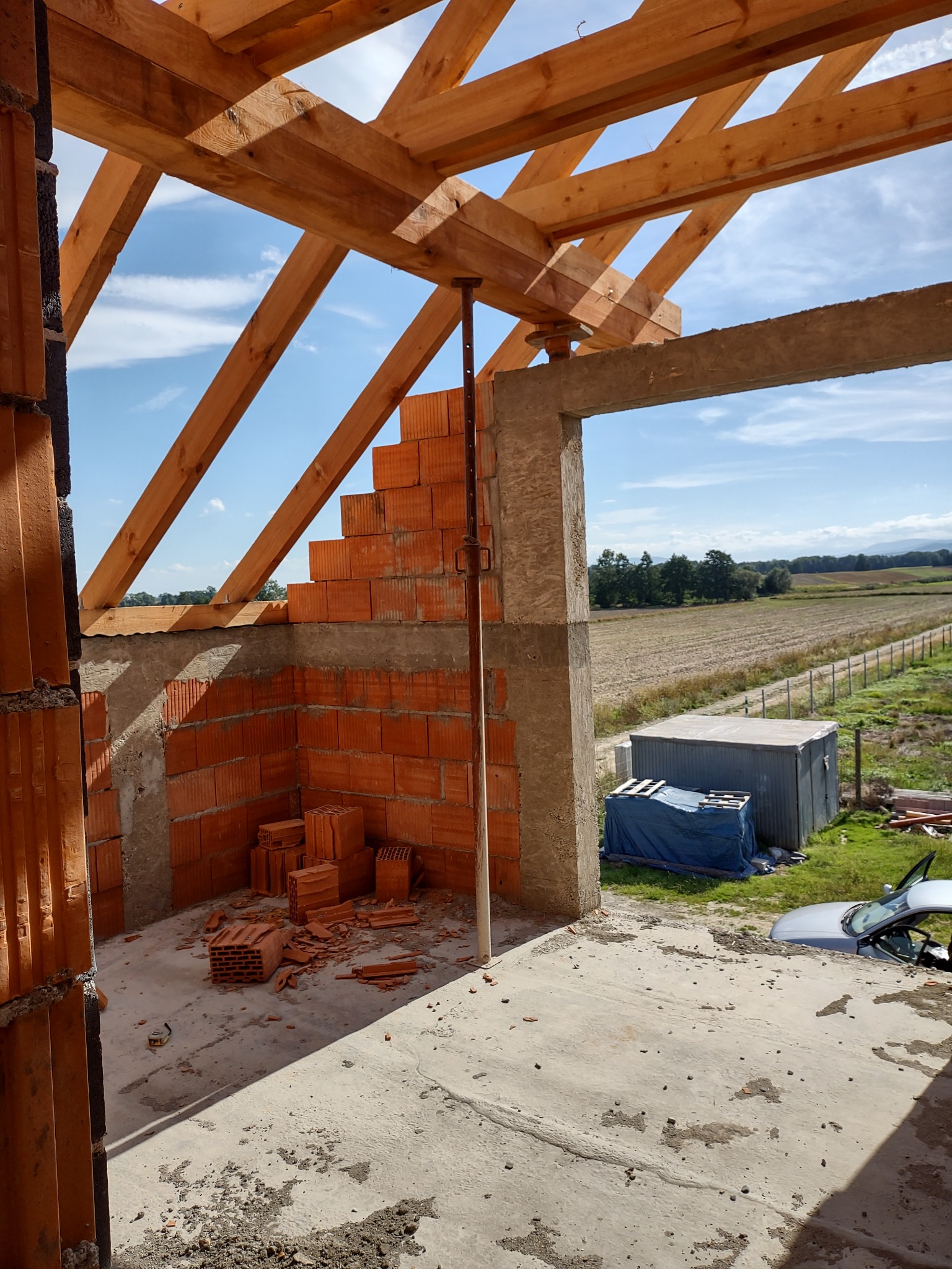 Surowa konstrukcja domu z cegły ceramicznej, widoczne drewniane krokwie dachu i podpora teleskopowa, w tle krajobraz z polem i niebem.