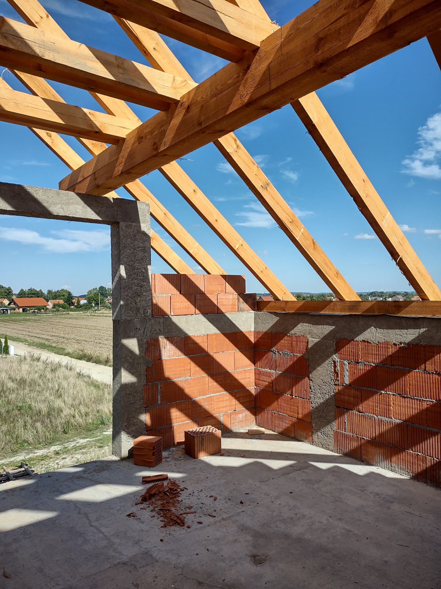 Surowa konstrukcja budynku w trakcie wznoszenia: widoczne ceglane ściany, betonowy nadproże i drewniana więźba dachowa na tle błękitnego nieba i wiejskiej zabudowy.