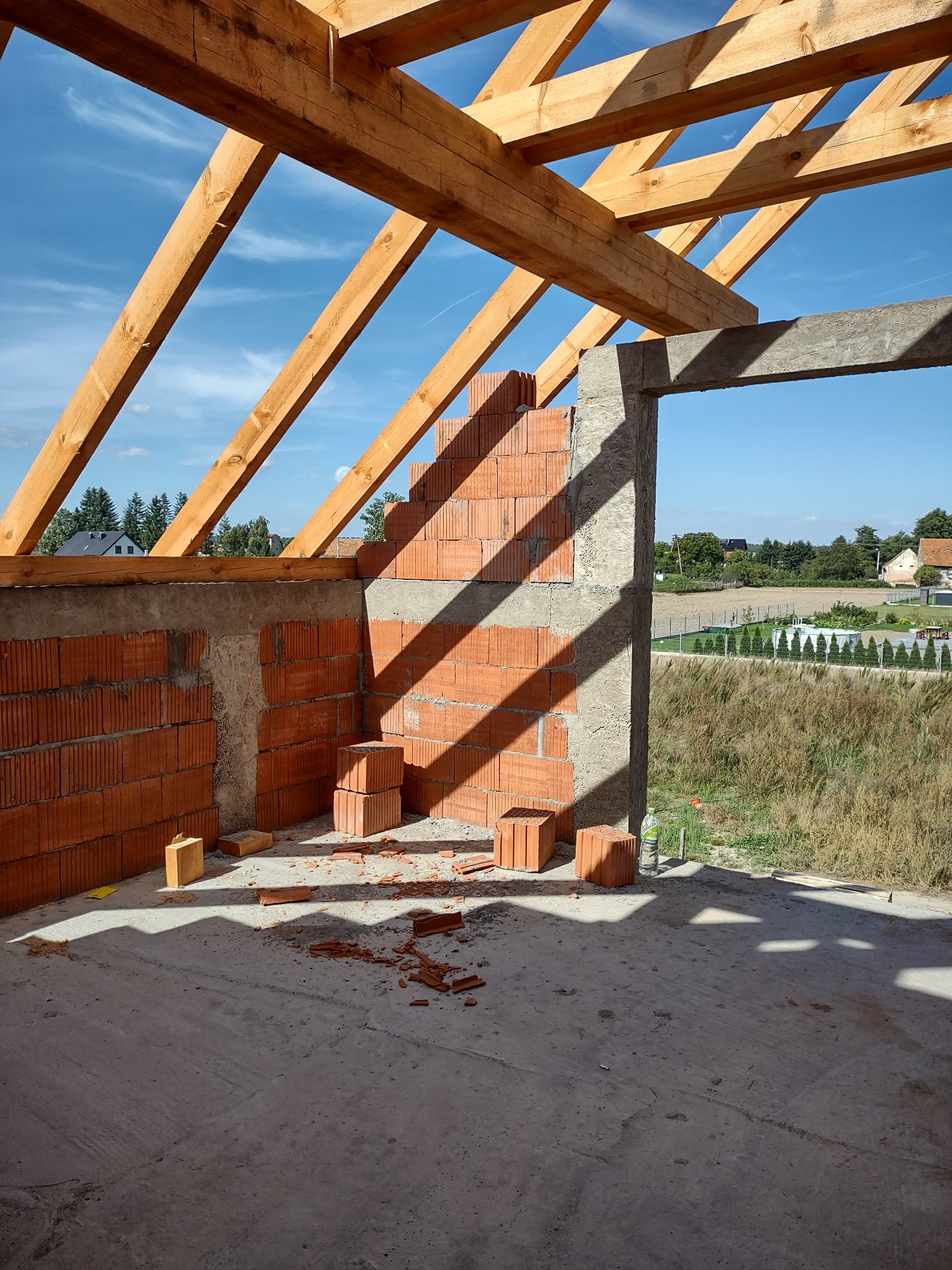 Surowa konstrukcja budynku w trakcie budowy: drewniany szkielet dachu, fragment ściany z czerwonej cegły, surowy betonowy otwór okienny z widokiem na wieś w słoneczny dzień.