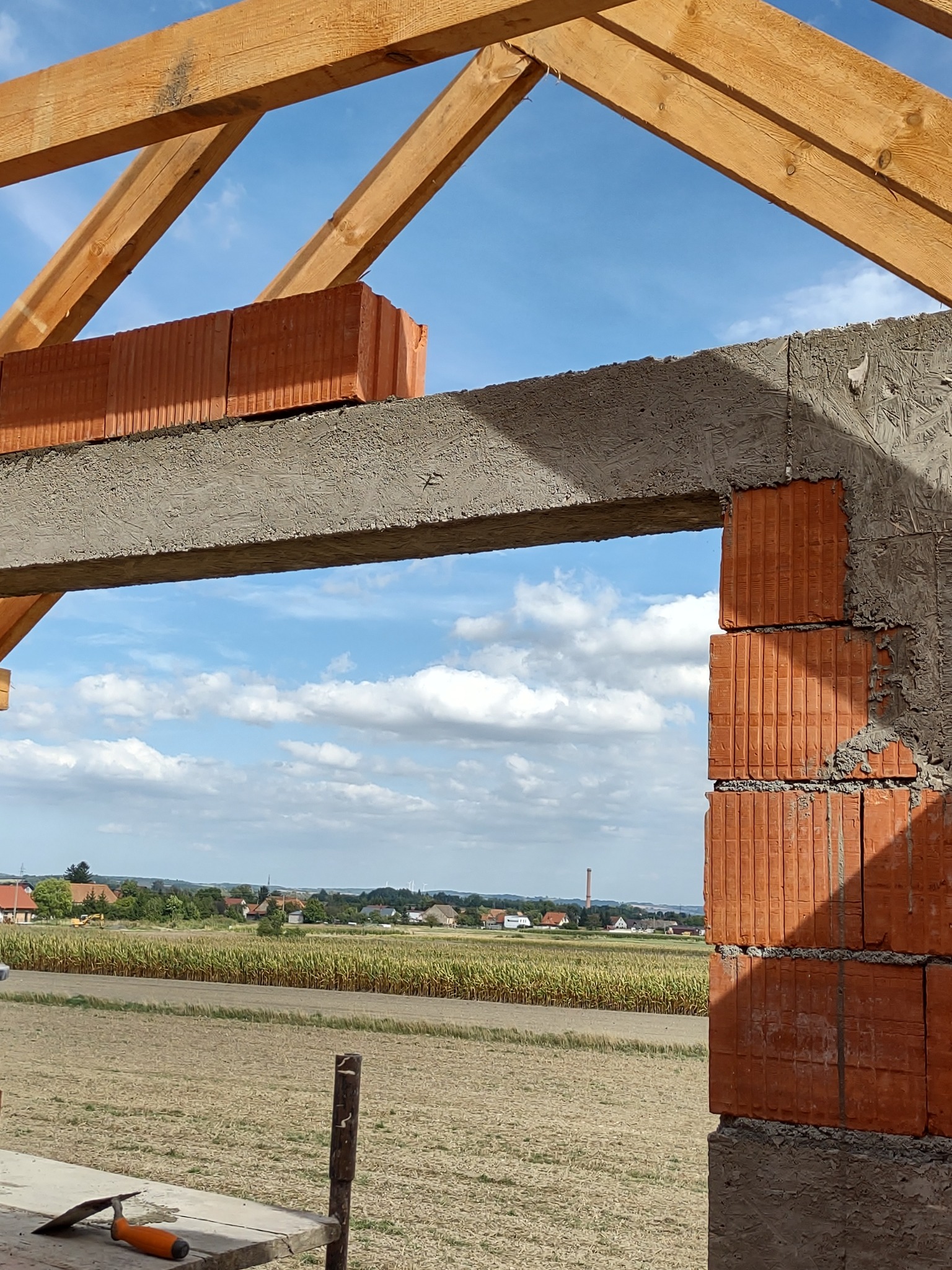 Wznoszenie muru z czerwonej cegły ceramicznej na budowie, widoczne drewniane elementy konstrukcyjne, narzędzie murarskie na pierwszym planie oraz krajobraz z polami i zabudowaniami w tle.