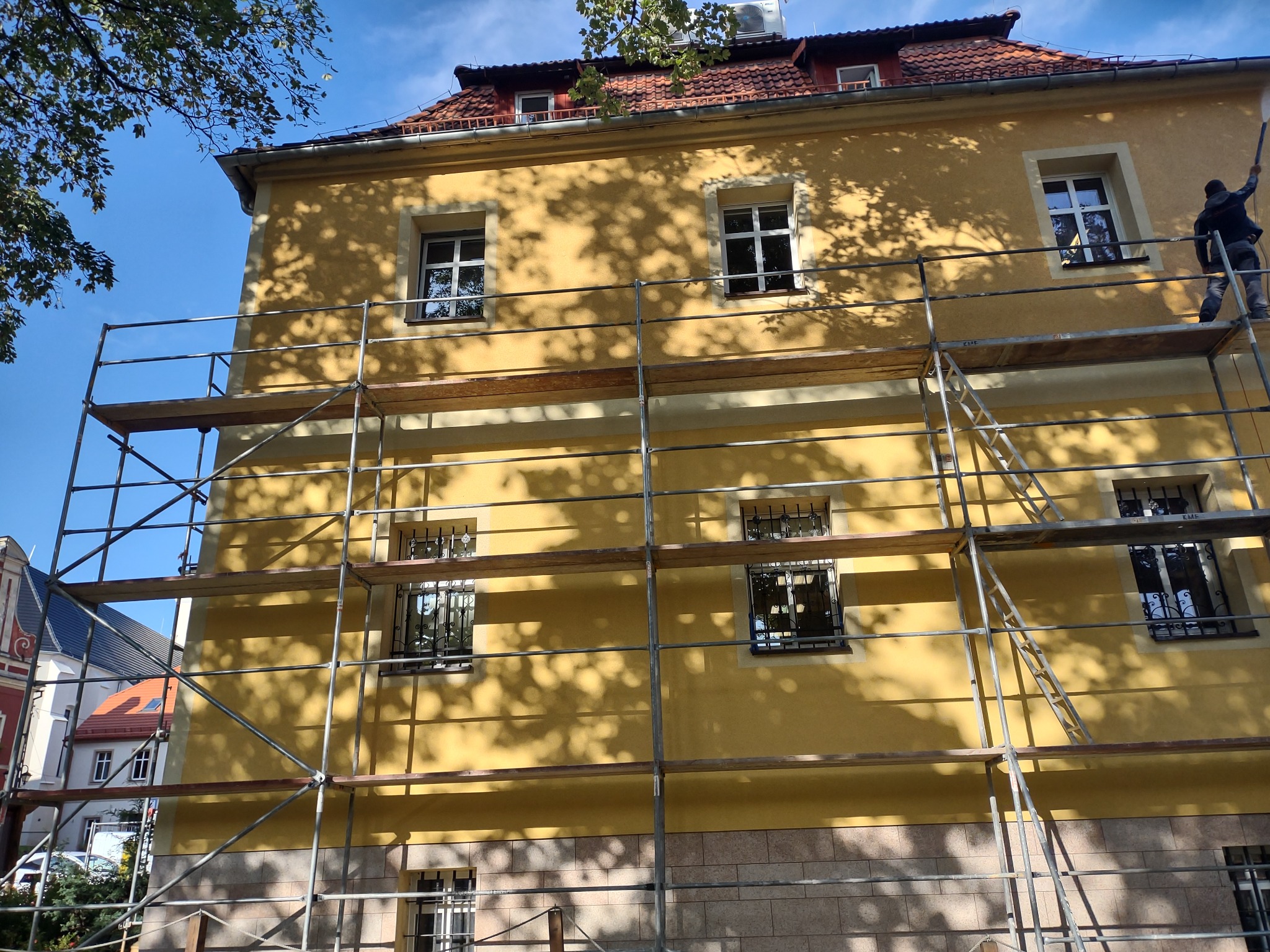 Żółta elewacja budynku w trakcie malowania, widoczne rusztowanie i pracownik z wałkiem na wysokości, rzucane cienie drzew na ścianie.