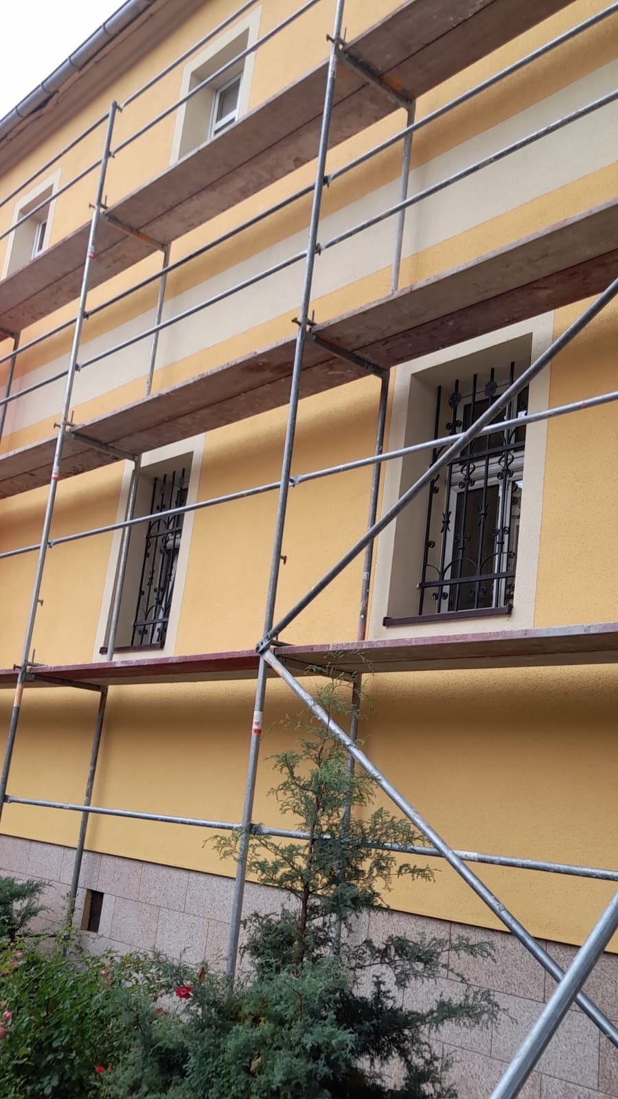 Żółta elewacja budynku w trakcie prac wykończeniowych, widoczne rusztowania, okna z ozdobnymi kratami i fragment ogrodu z krzewami.