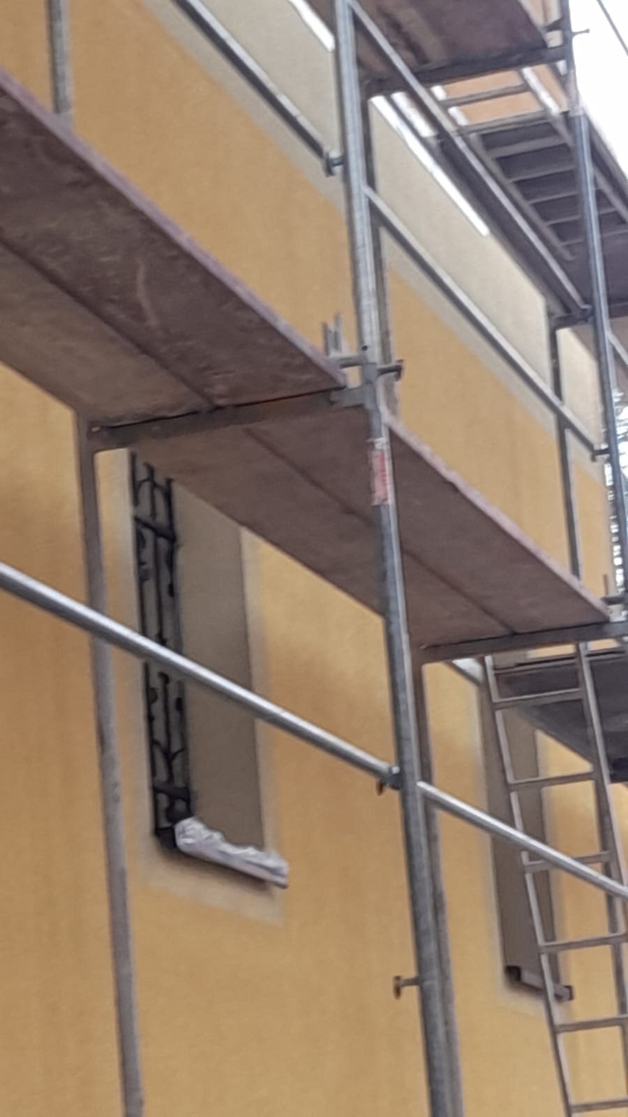 Fragment żółtej elewacji budynku w trakcie prac, widoczne rusztowanie z drewnianymi podestami, zabezpieczone okno folią malarską.