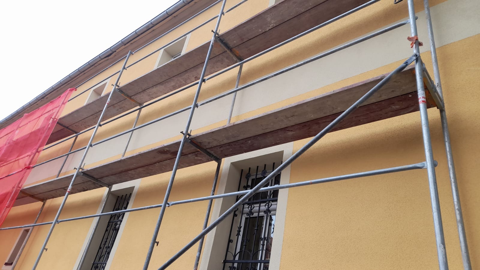 Żółta elewacja budynku w trakcie prac wykończeniowych, widoczne rusztowania z czerwonym zabezpieczeniem siatkowym, okna z ozdobnymi kratami.