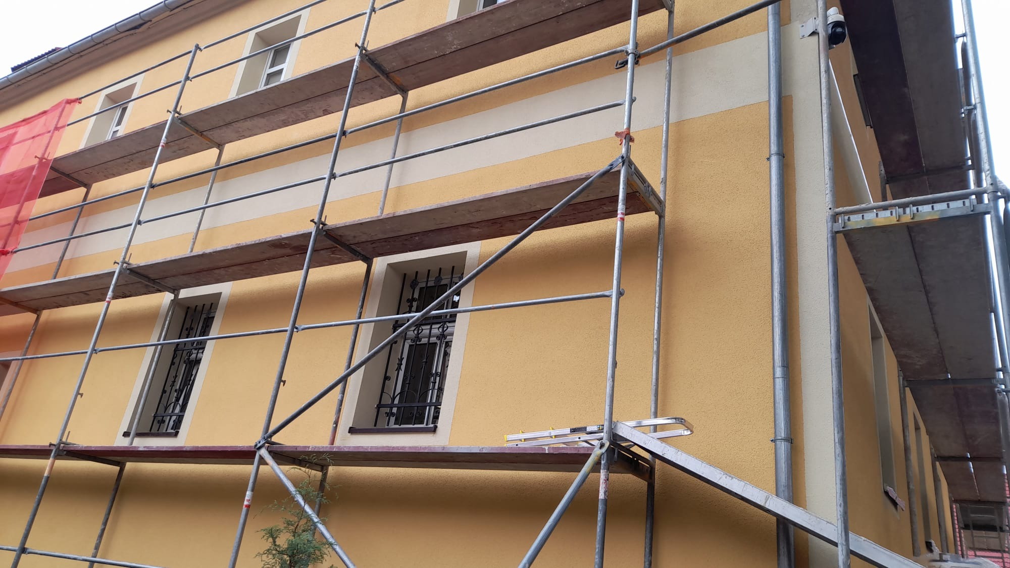 Żółta elewacja budynku w trakcie prac, widoczne rusztowania i fragment siatki zabezpieczającej, z zamontowaną kamerą monitoringu na rogu budynku.