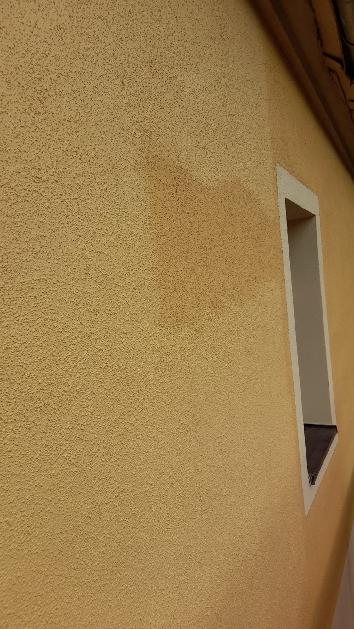 Fragment żółtej elewacji z wyraźną plamą wilgoci i wnęką okienną z białym obramowaniem.