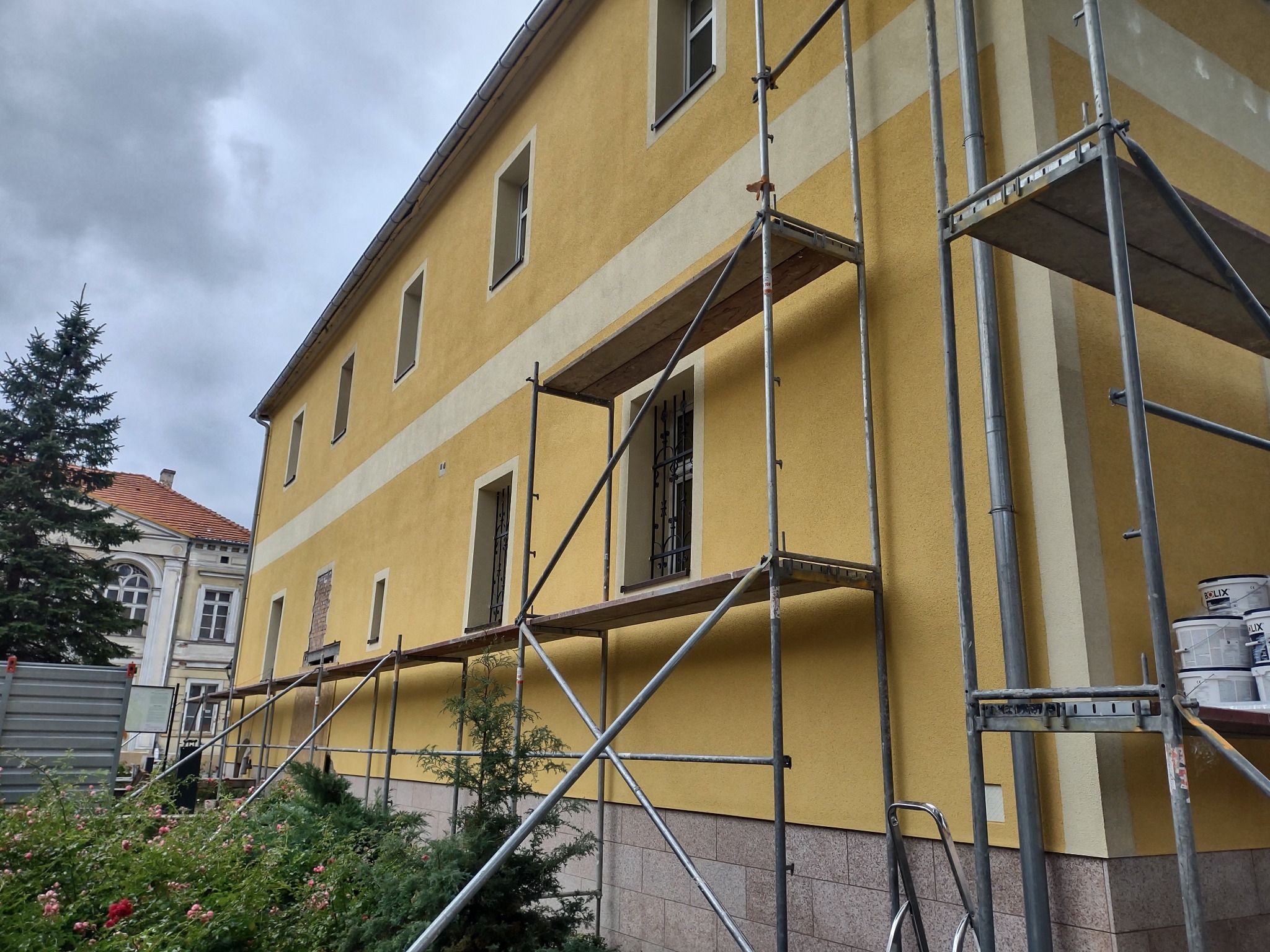 Żółta elewacja budynku z jasnym pasem, w trakcie malowania, widoczne rusztowania, puszki z farbą Bolix i fragment sąsiedniego budynku z czerwonym dachem.