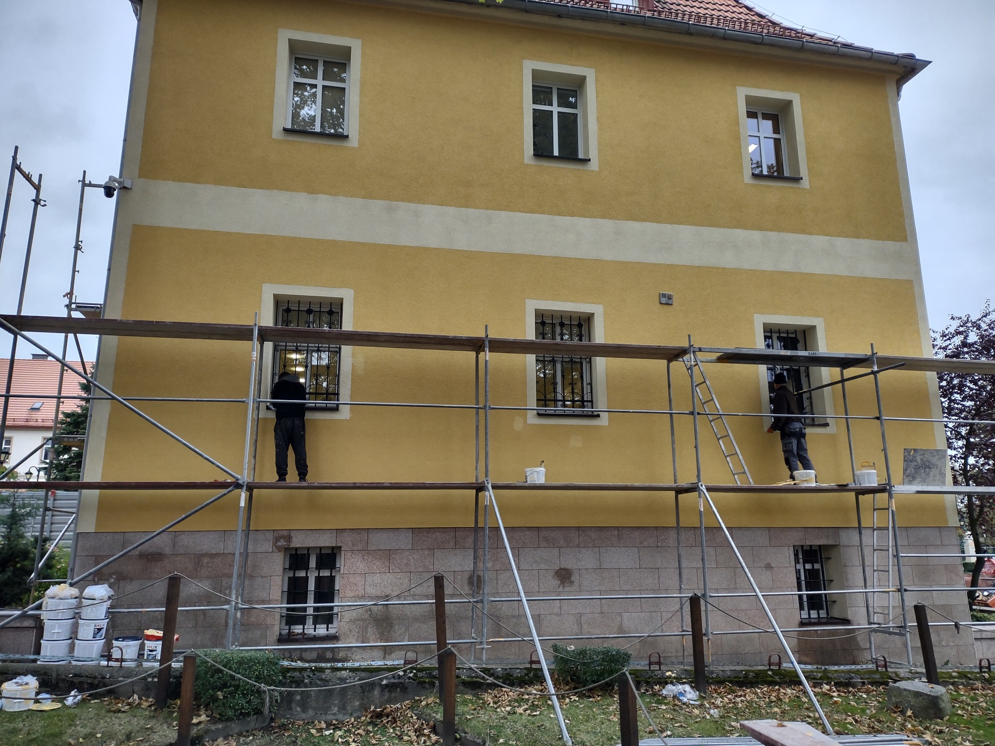 Dwóch pracowników na rusztowaniu maluje elewację dwukondygnacyjnego budynku w odcieniach żółci i beżu; widoczne wiadra z farbą i narzędzia pracy.