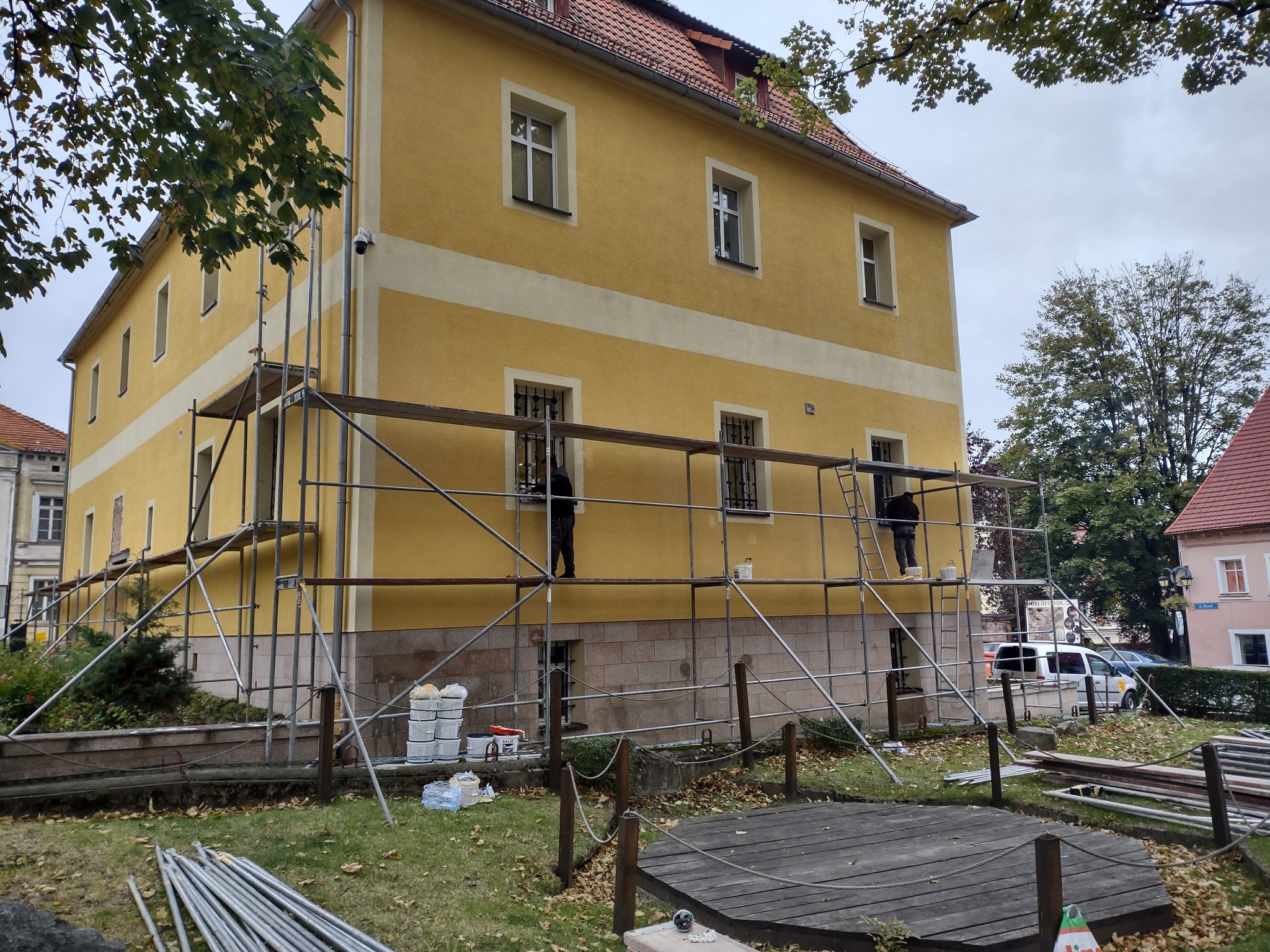 Żółty budynek w trakcie renowacji elewacji, widoczne rusztowania, pracownicy oraz puszki z farbą, na pierwszym planie elementy drewnianej konstrukcji.