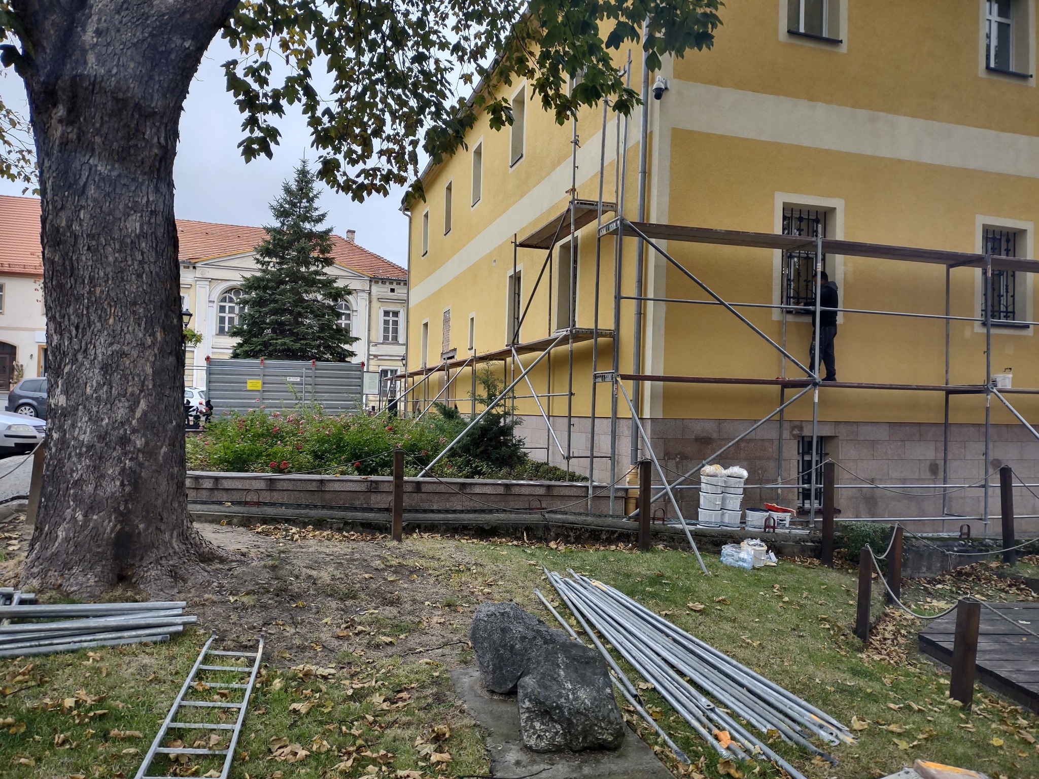 Pracownik w ciemnej odzieży maluje elewację żółtego budynku z białymi pasami, stojąc na rusztowaniu. Na pierwszym planie leżą rury rusztowania, drabina i dwa duże kamienie, a w tle widać inne...