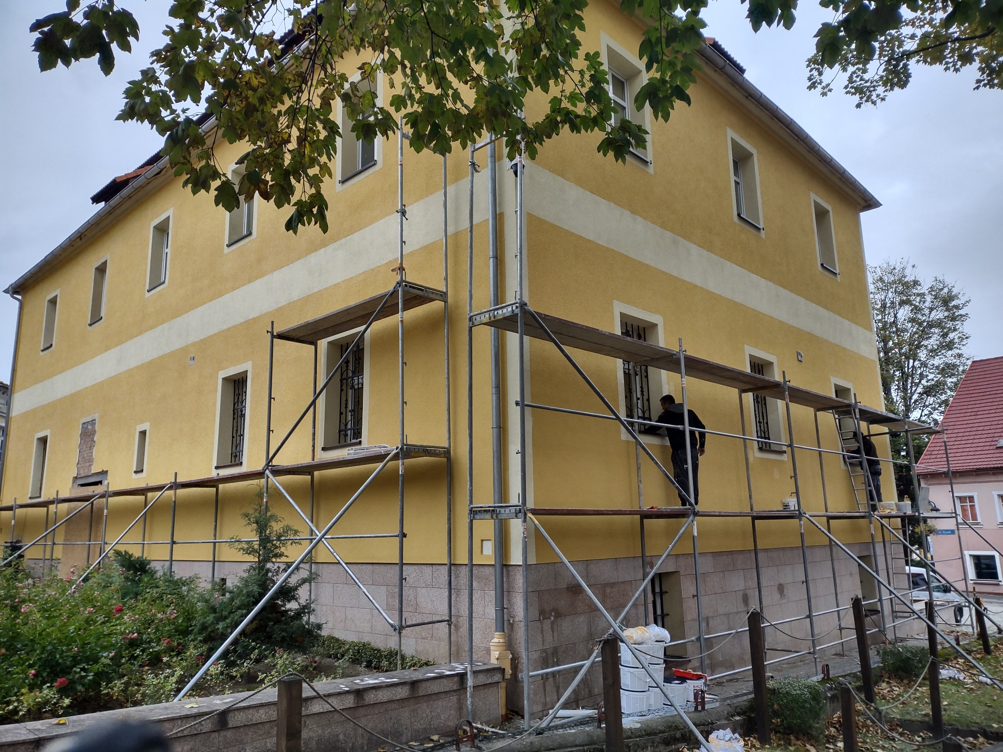Żółta elewacja dwupiętrowego budynku w Lądku-Zdroju podczas prac renowacyjnych, z widocznym fragmentem usuniętego tynku i rusztowaniami, na których pracują dwie osoby.