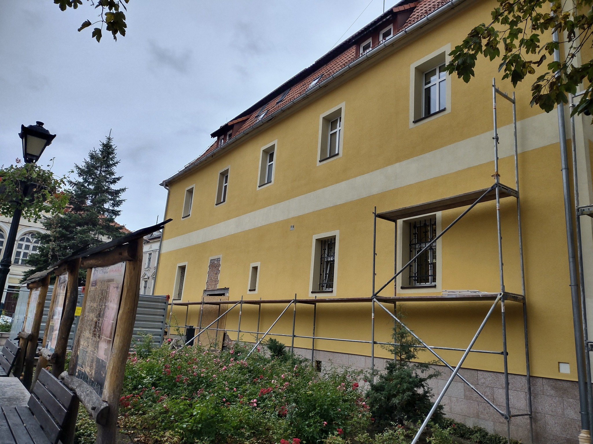 Żółta elewacja budynku w trakcie prac, widoczne rusztowania i fragmenty odsłoniętej cegły, na pierwszym planie rabata z różami.