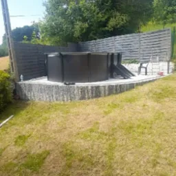 Czarny, okrągły basen ogrodowy z drabinką i krzesłem na kamiennym podwyższeniu, otoczony szarym, drewnianym ogrodzeniem w Zawierciu.