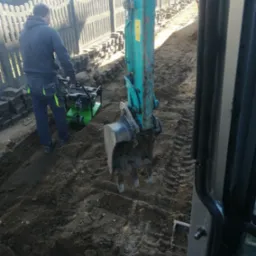 Prace ziemne: operator zagęszczarki w pobliżu wykopu pod ogrodzenie z prefabrykatów betonowych, widoczna łyżka koparki i ślady opon.