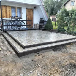 Dwu-poziomowy taras z kostki brukowej w trakcie budowy, z widocznymi elementami wykończeniowymi i przygotowanym podłożem, na tle domu z drewnianą elewacją i rowerami na tarasie.