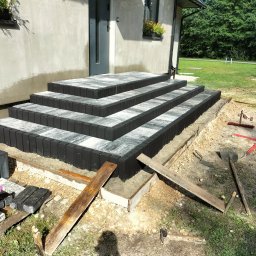 Usługi Brukarskie - Schody wejściowe z kostki brukowej w trakcie budowy, widoczne narzędzia i drewniane szalunki, dom z oknami i kwiatami w tle.