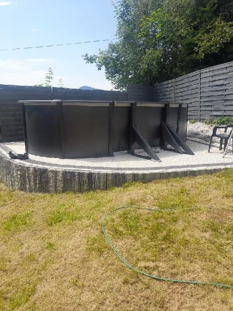 Częściowo wkopany, czarny basen ogrodowy z drewnianą obudową, otoczony białym żwirem i obrzeżem z betonowych elementów, na tle drewnianego ogrodzenia i trawnika.