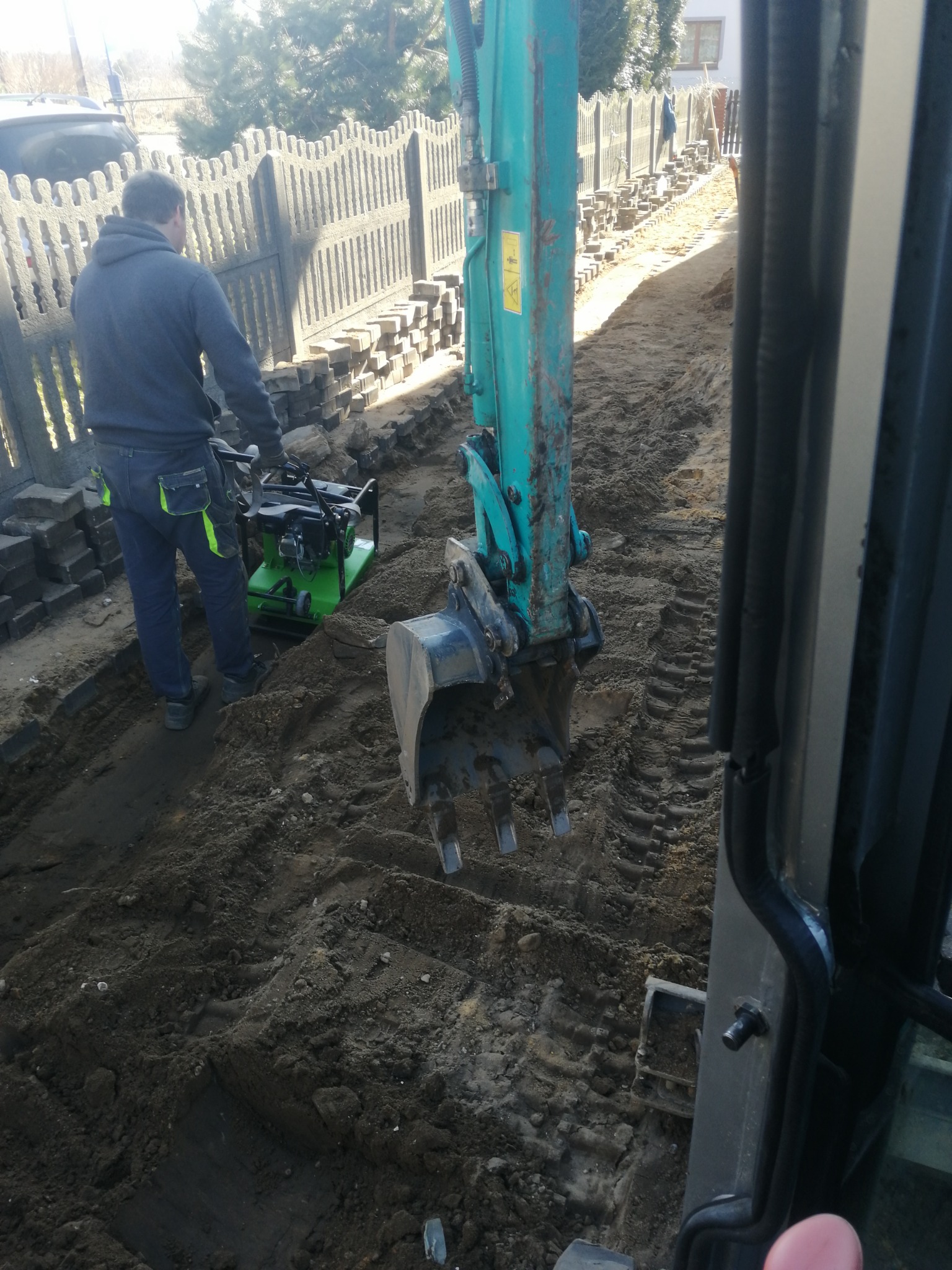 Prace ziemne: operator zagęszczarki w pobliżu wykopu pod ogrodzenie z prefabrykatów betonowych, widoczna łyżka koparki i ślady opon.