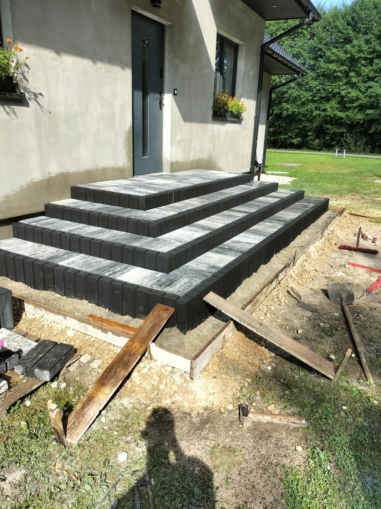 Schody wejściowe z kostki brukowej w trakcie budowy, widoczne narzędzia i drewniane szalunki, dom z oknami i kwiatami w tle.