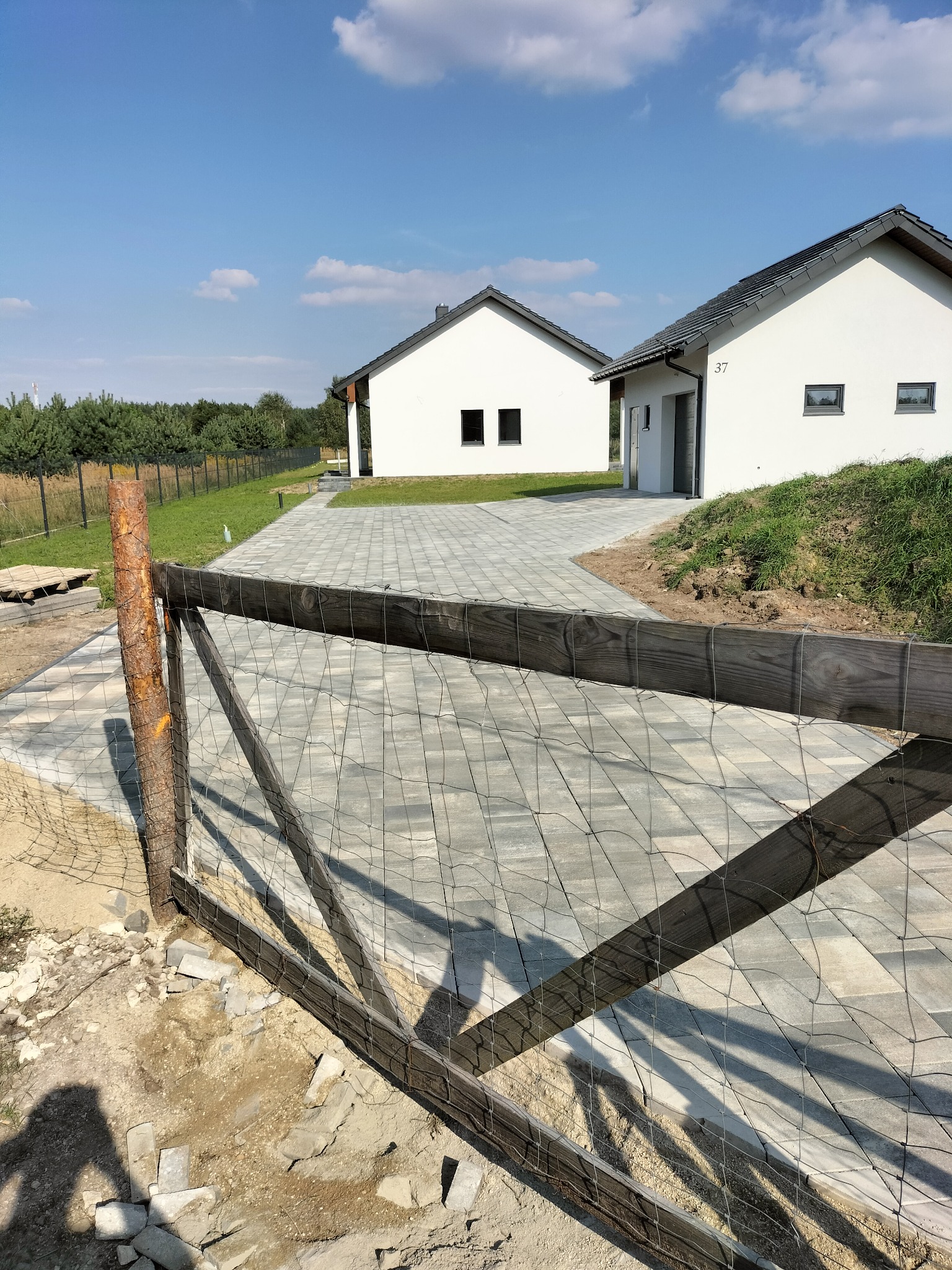 Surowy drewniany płot z siatką na tle świeżo ułożonej kostki brukowej prowadzącej do dwóch białych domów z szarymi dachami w słoneczny dzień.
