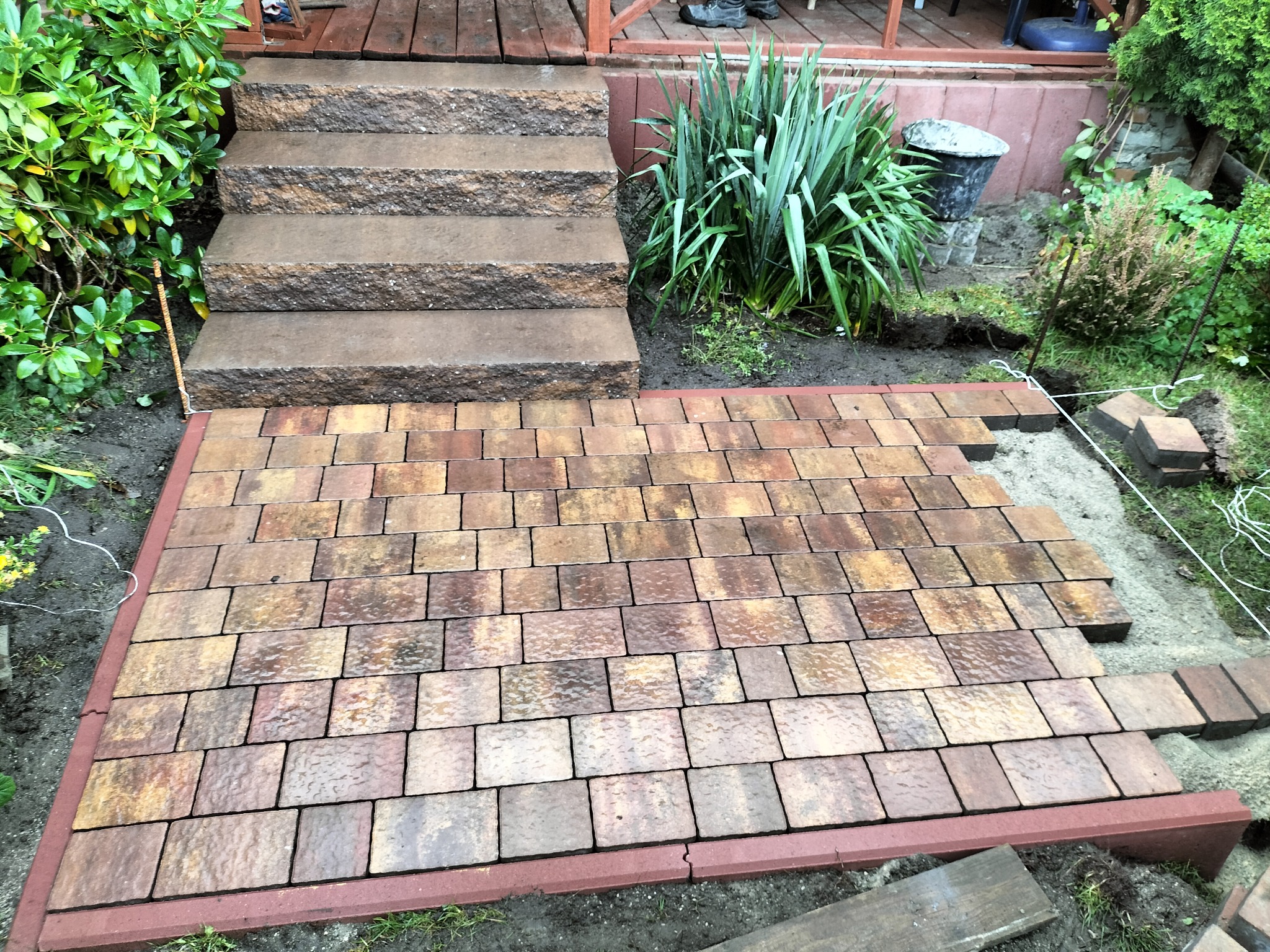 Układanie brązowo-beżowej kostki brukowej na przygotowanym podłożu, widoczne schody kamienne prowadzące do drewnianego tarasu, roślinność w tle i częściowo ułożone obrzeże.