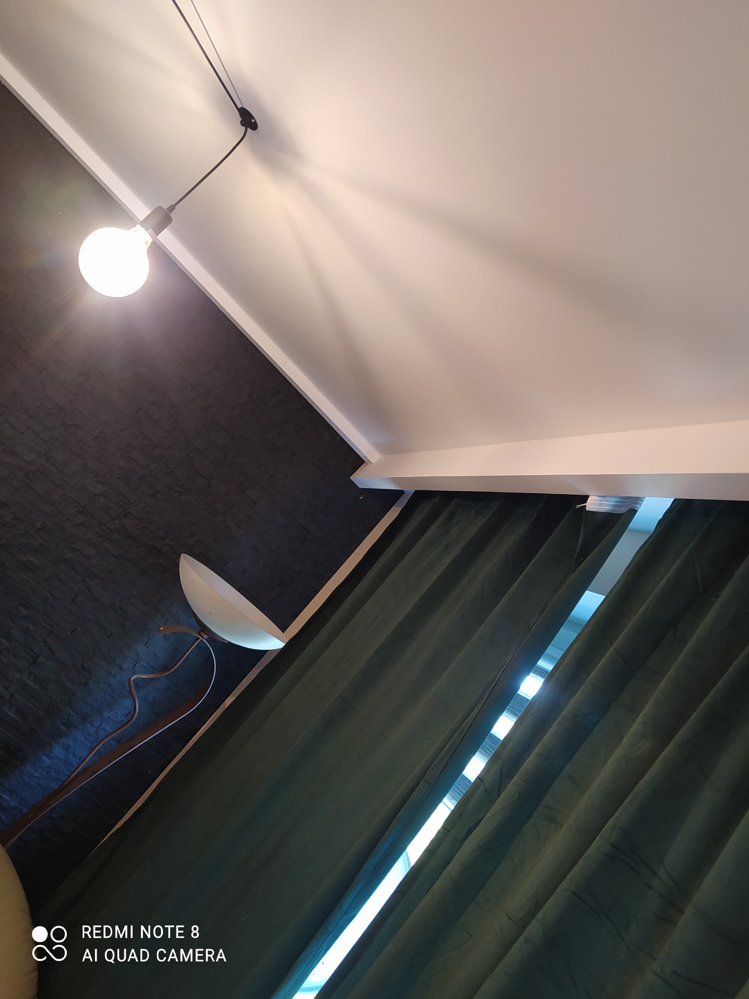 Ciemna ściana z fakturą kamienia, lampa wisząca z żarówką Edison, zielone zasłony z prześwitem okna, biały sufit, nietypowy kąt ujęcia.