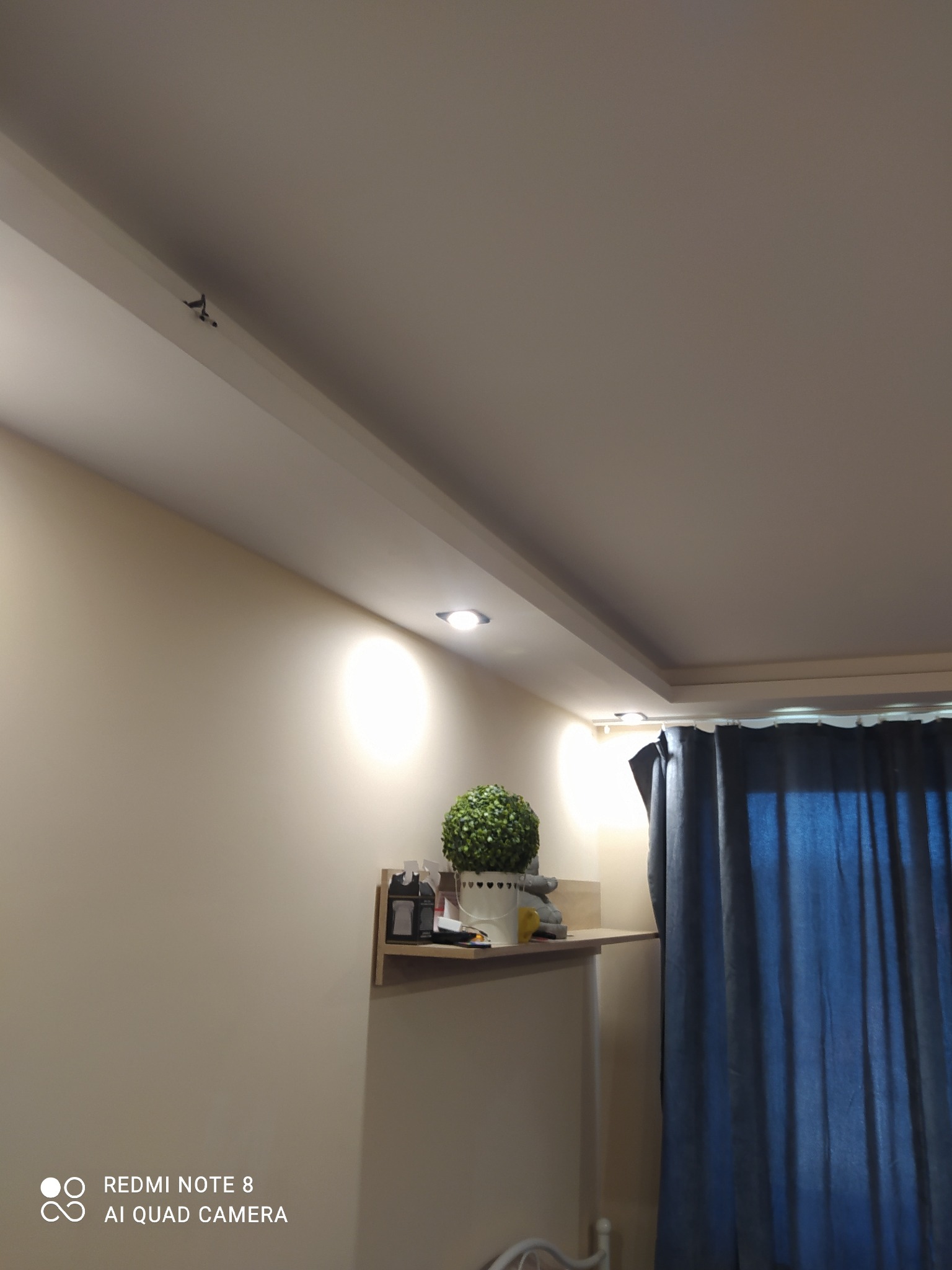 Wykończony sufit podwieszany z oświetleniem punktowym w pokoju, widoczna półka z dekoracjami i zasłony.