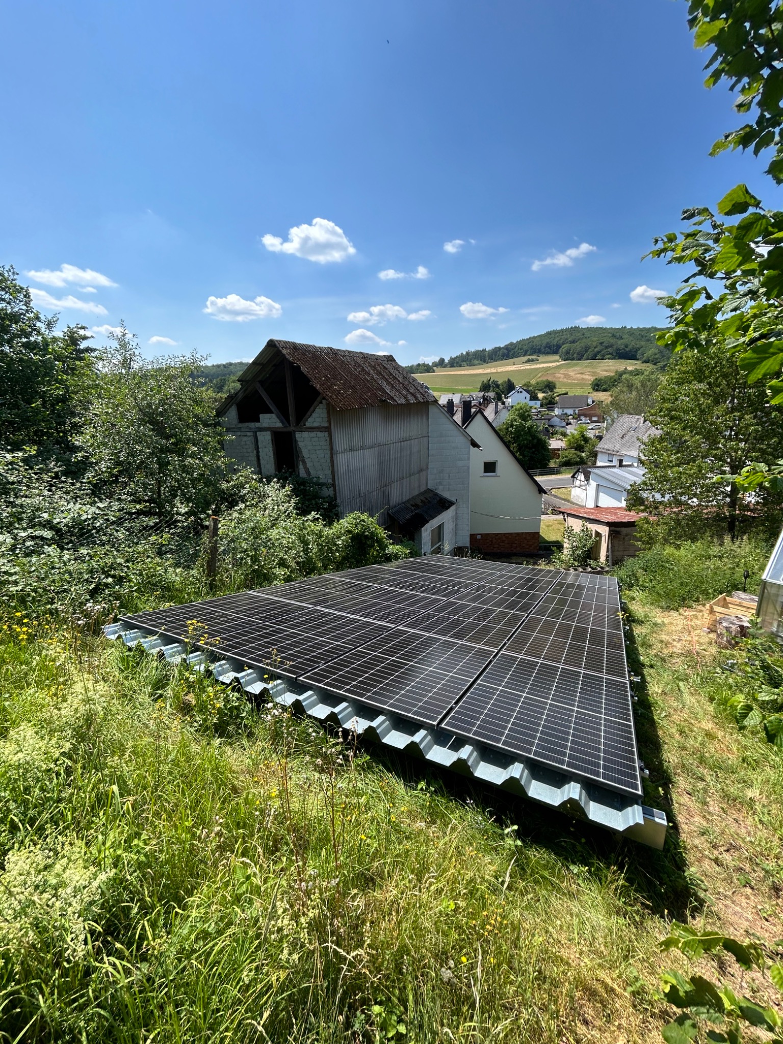 Panel słoneczny na falistym dachu w wiejskim otoczeniu, z widokiem na starą stodołę i okoliczne domy pod błękitnym niebem.