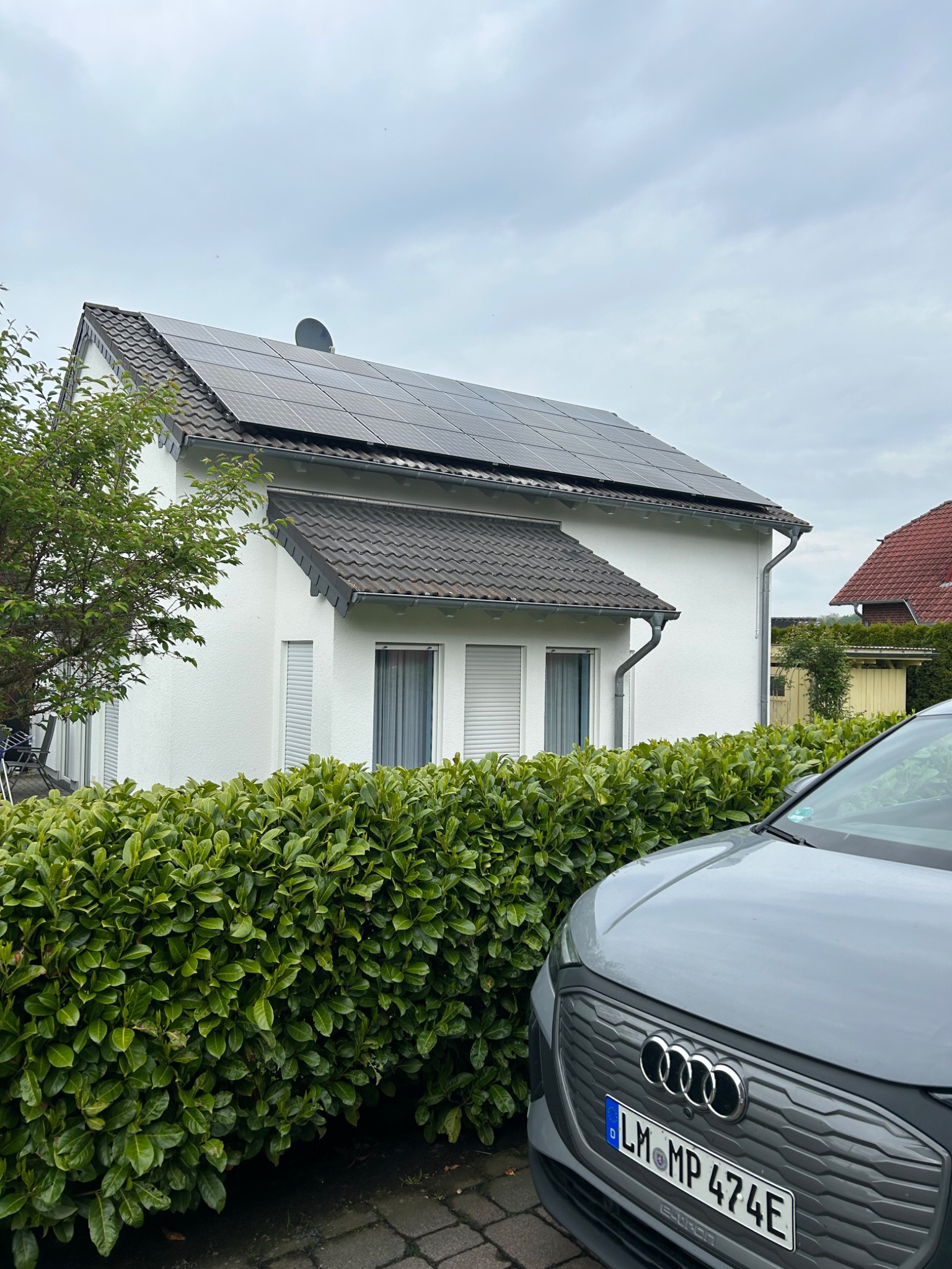 Biały dom jednorodzinny z panelami słonecznymi na dachu, widoczny szary samochód elektryczny Audi i zadbany żywopłot.