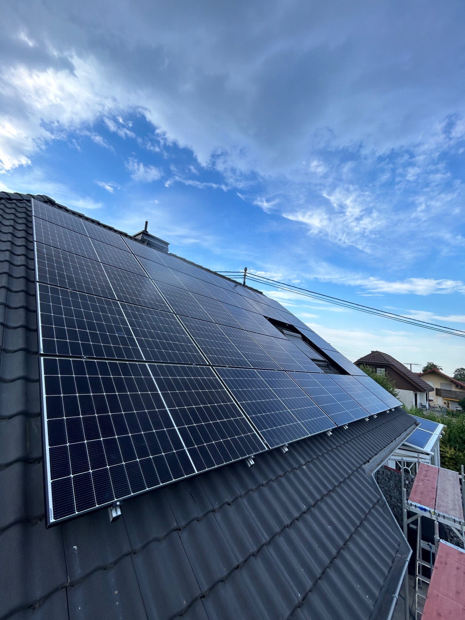 Panele słoneczne na ciemnym dachu z dachówki ceramicznej, widoczne rusztowanie z boku i fragment niebieskiego nieba z chmurami. Jeden z paneli jest częściowo otwarty, ukazując mechanizm.