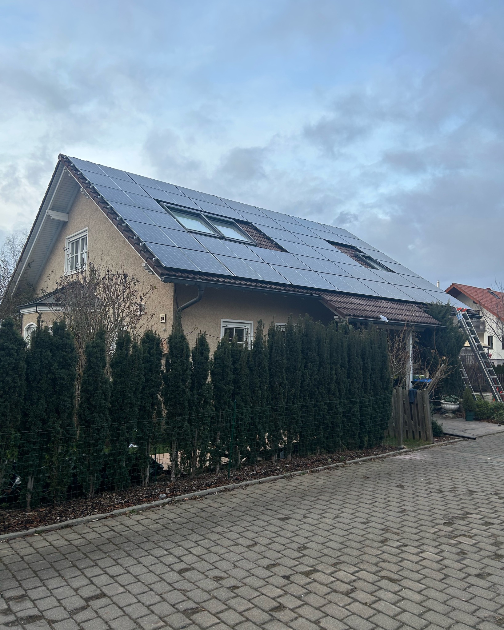 Dom jednorodzinny z panelami słonecznymi na dachu, widoczne okna dachowe i drabina oparta o ścianę sąsiedniego budynku.