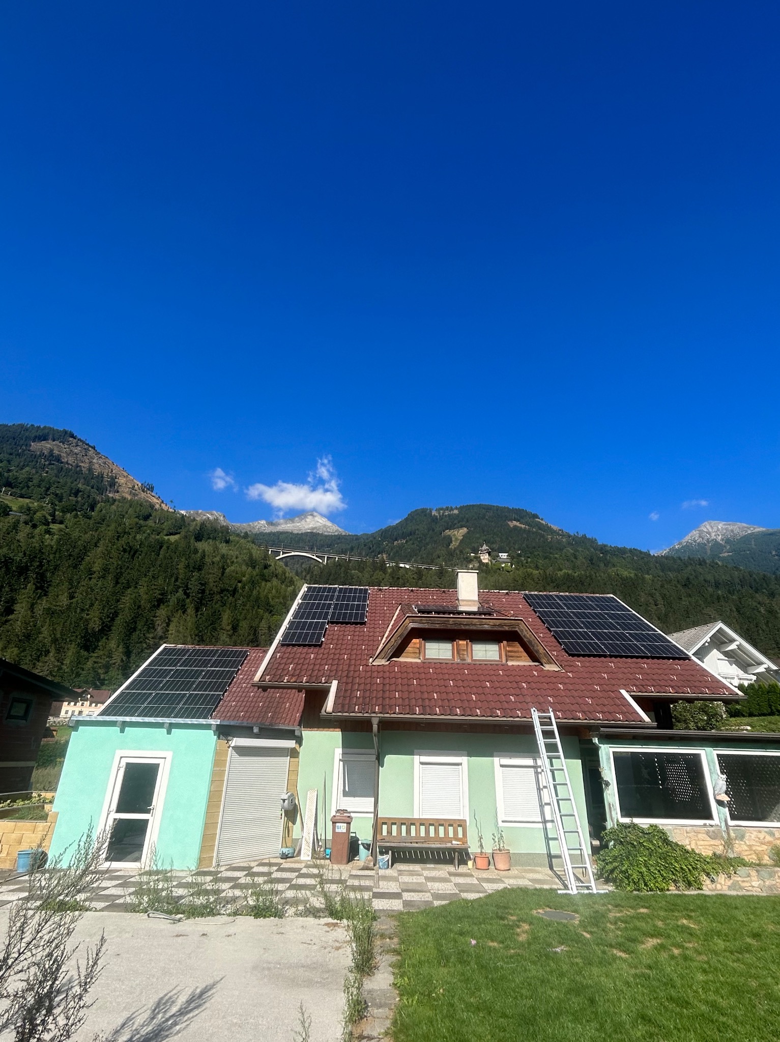 Dom z czerwoną dachówką i panelami słonecznymi, drabina oparta o ścianę, w tle góry i most.