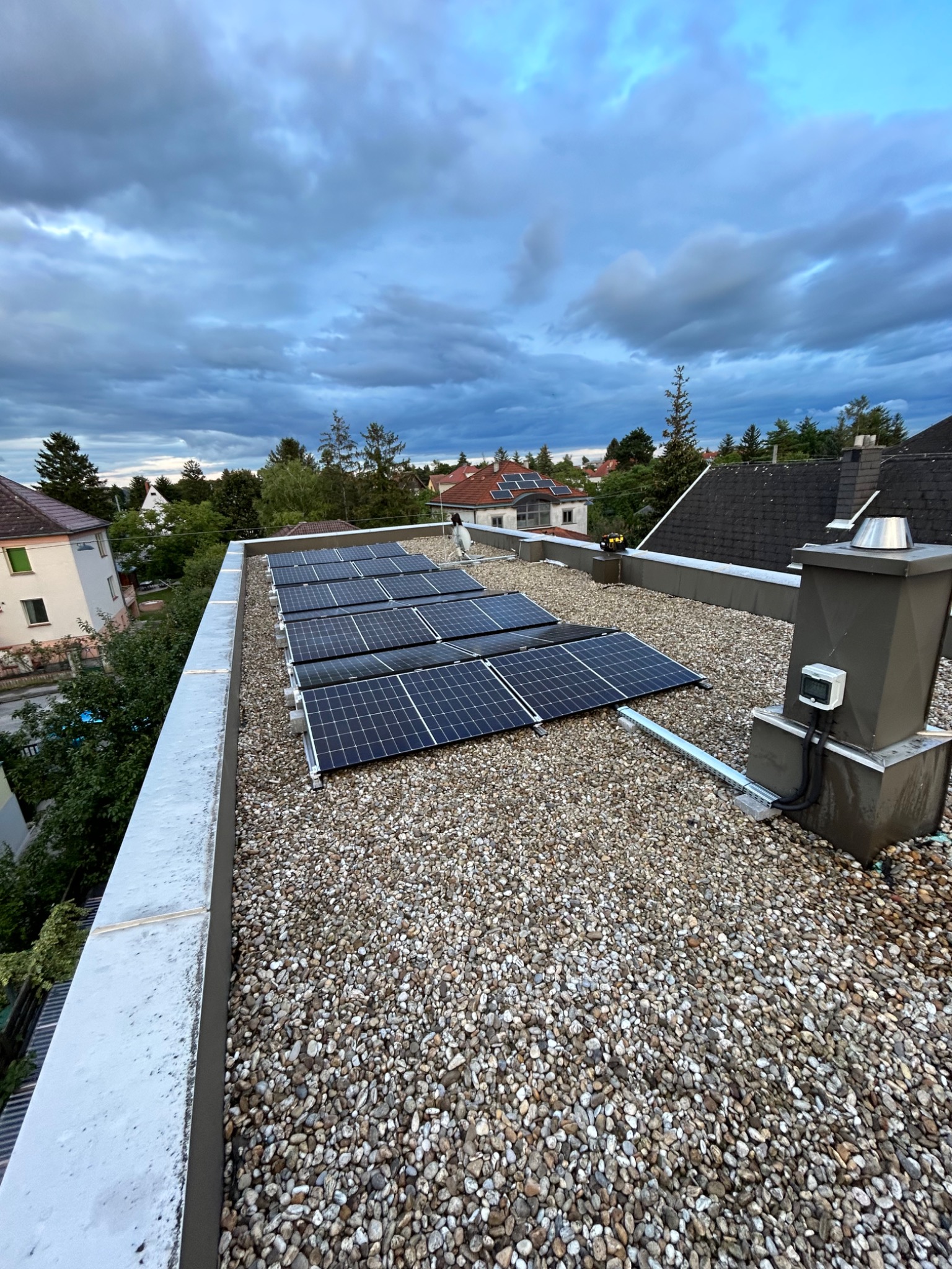 Panele słoneczne zamontowane na płaskim dachu pokrytym drobnym żwirem, widok z góry na tle pochmurnego nieba i zabudowań.