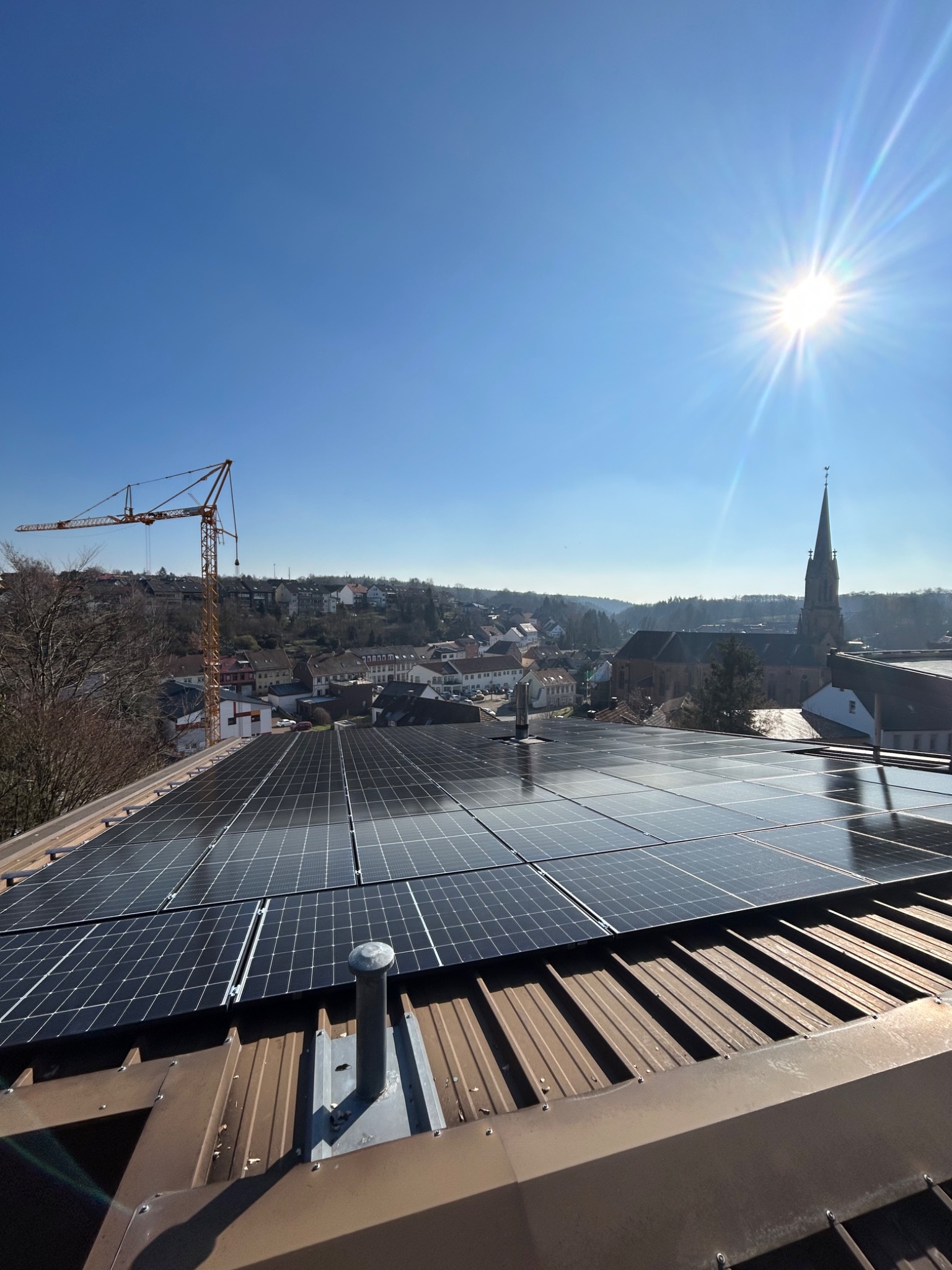 Panel słoneczny na brązowym dachu z widokiem na miasto i kościół w słoneczny dzień, z żurawiem budowlanym w tle.