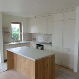 Jasna kuchnia z drewnianym blatem wyspy i złotą armaturą, widoczne niedokończone podłogi i wystające przewody z sufitu.