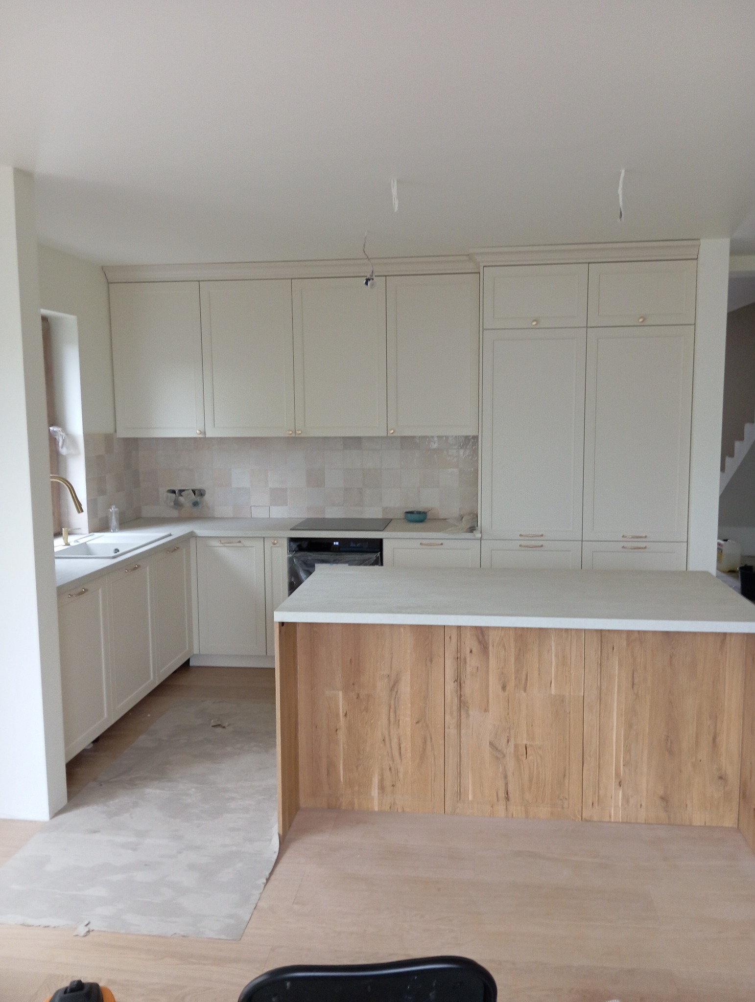 Jasna kuchnia z drewnianą wyspą w trakcie montażu, widoczne zabezpieczenia podłogi i wiszące kable oświetleniowe.