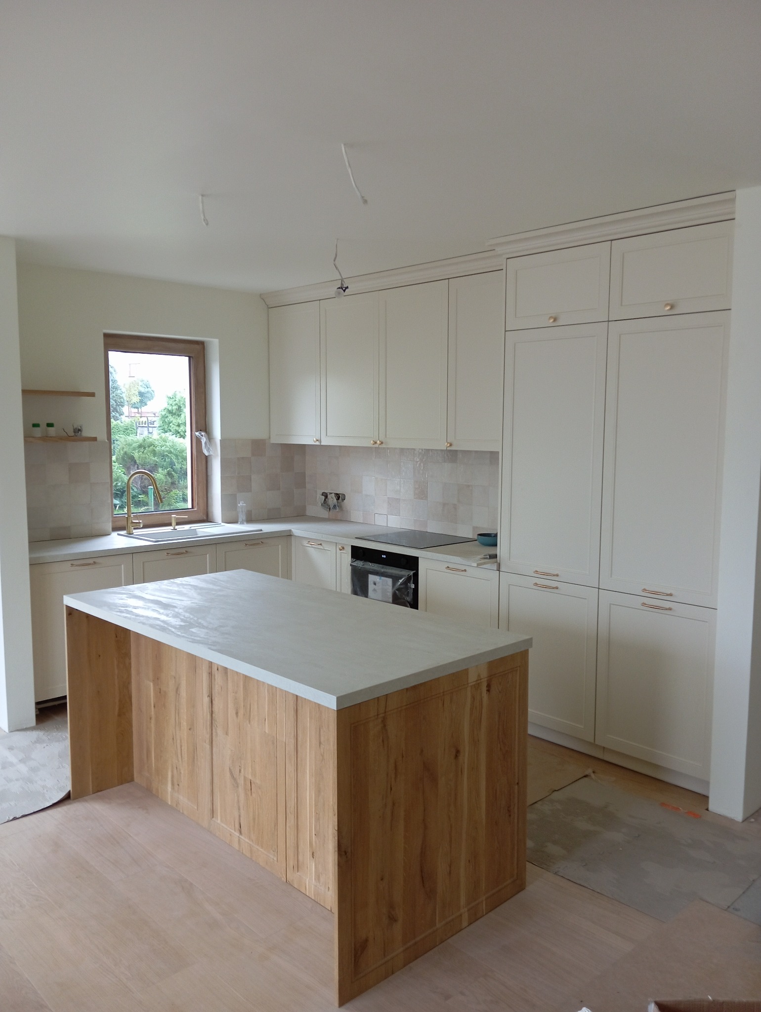 Jasna kuchnia z drewnianym blatem wyspy i złotą armaturą, widoczne niedokończone podłogi i wystające przewody z sufitu.