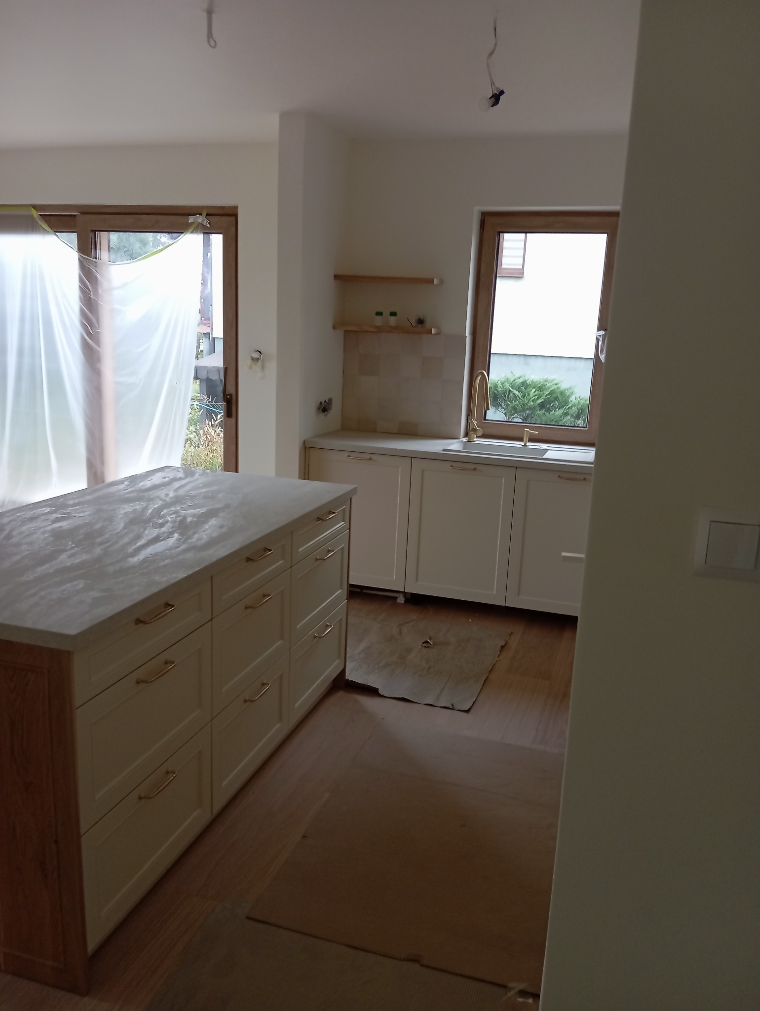 Wnętrze kuchni w trakcie montażu, widoczna wyspa z marmurowym blatem, białe szafki i zabezpieczona folią podłoga.